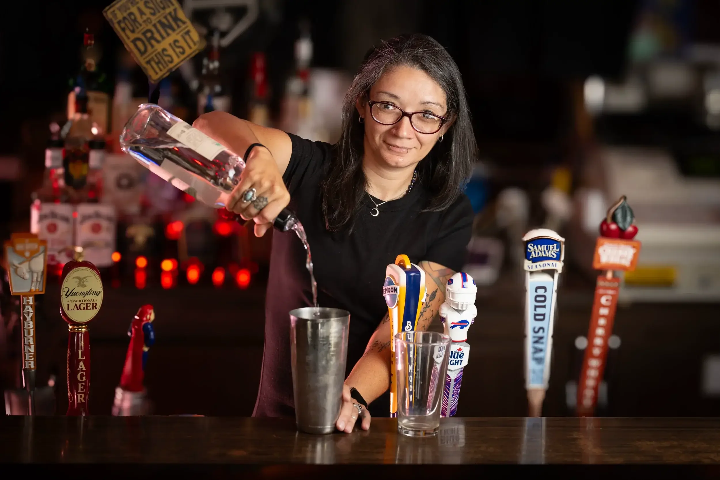 A woman with glasses and tattoos pouring a clear liquid into a glass at a bar with various beer tap handles and liquor bottles in the background.