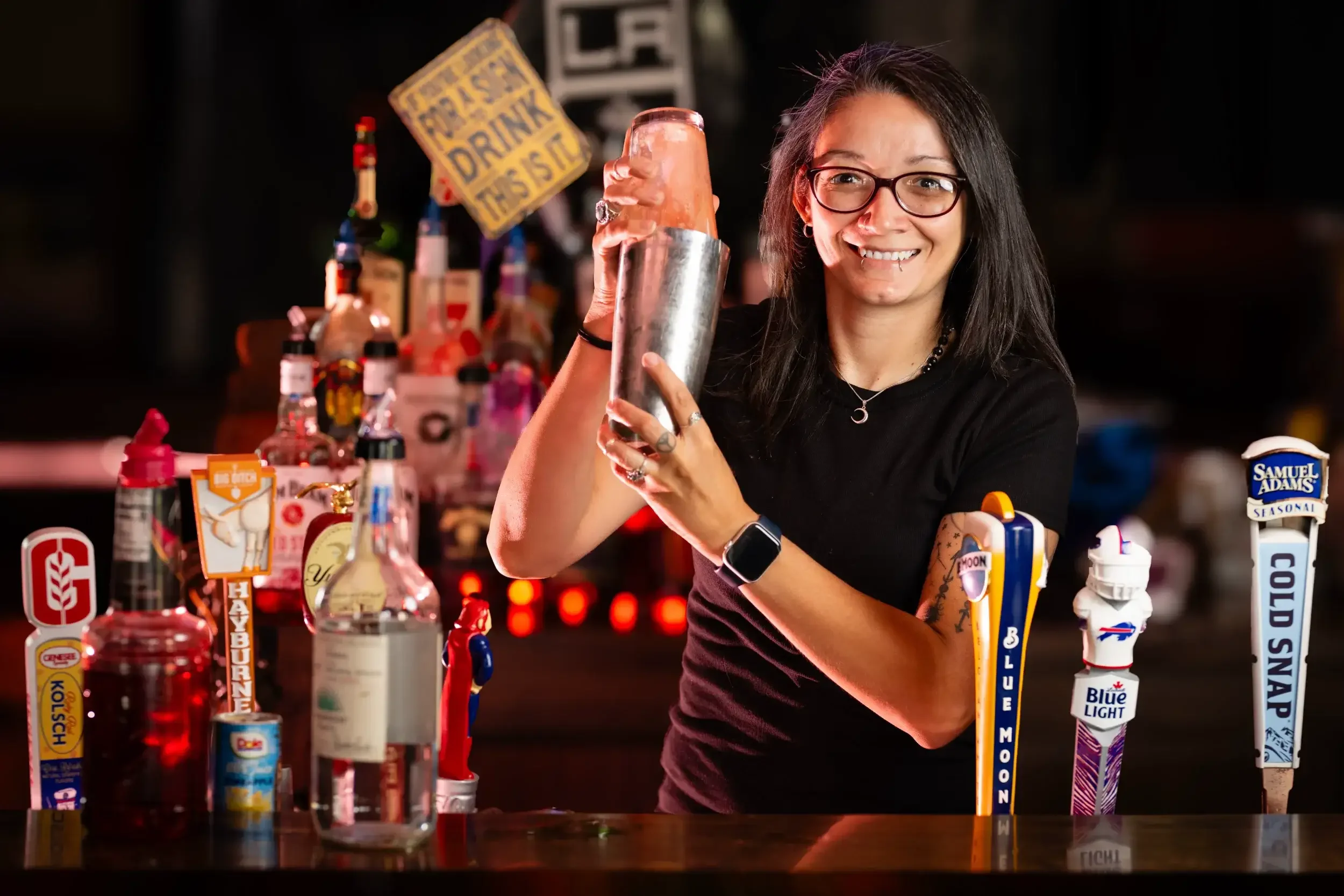 A woman behind a bar counter shaking a cocktail shaker, with assorted bottles of alcohol and drink garnishes visible around her.