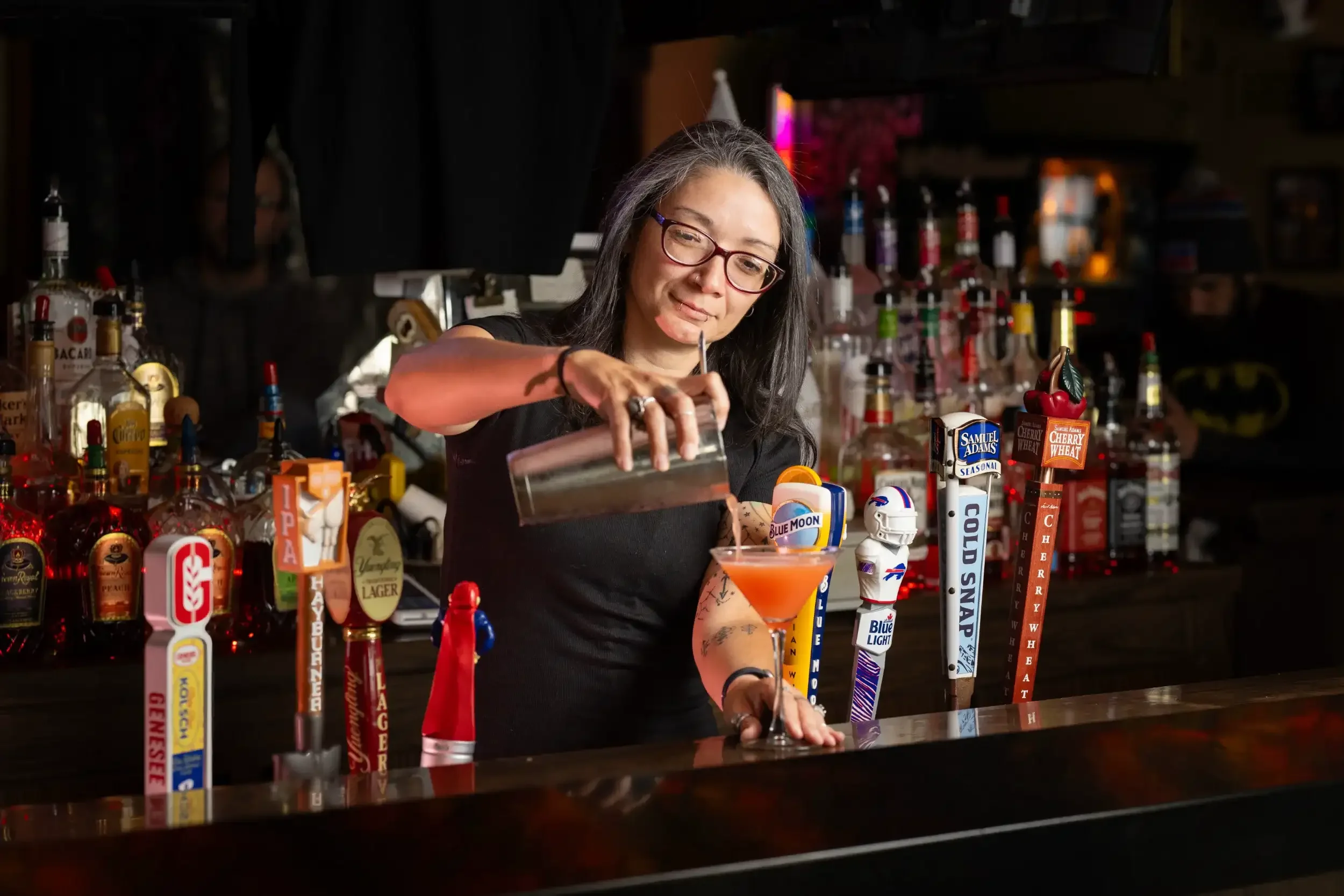 A woman with long gray hair and glasses is preparing a cocktail behind a bar. The bar is decorated with various liquor bottles and beer taps, including brands like Samuel Adams and Blue Moon. She is pouring a pink-colored drink into a glass with a cherry garnish.