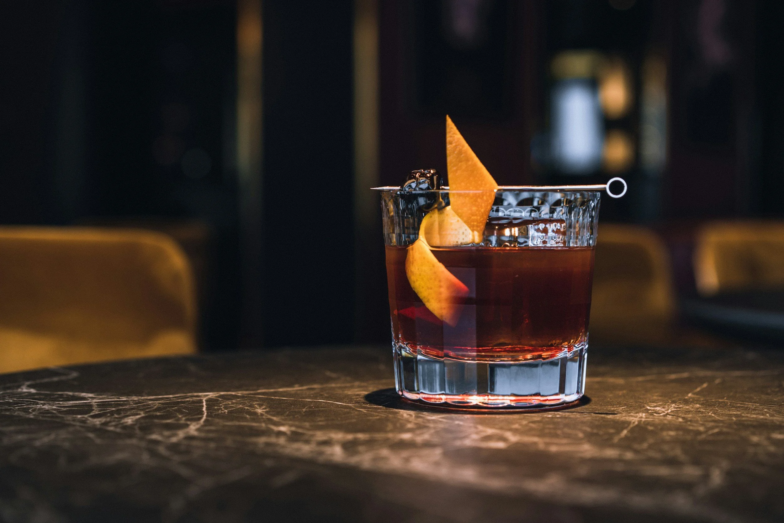 A close-up of a glass filled with a dark red cocktail, garnished with a lemon peel and a small black garnish, sitting on a dark marble surface.