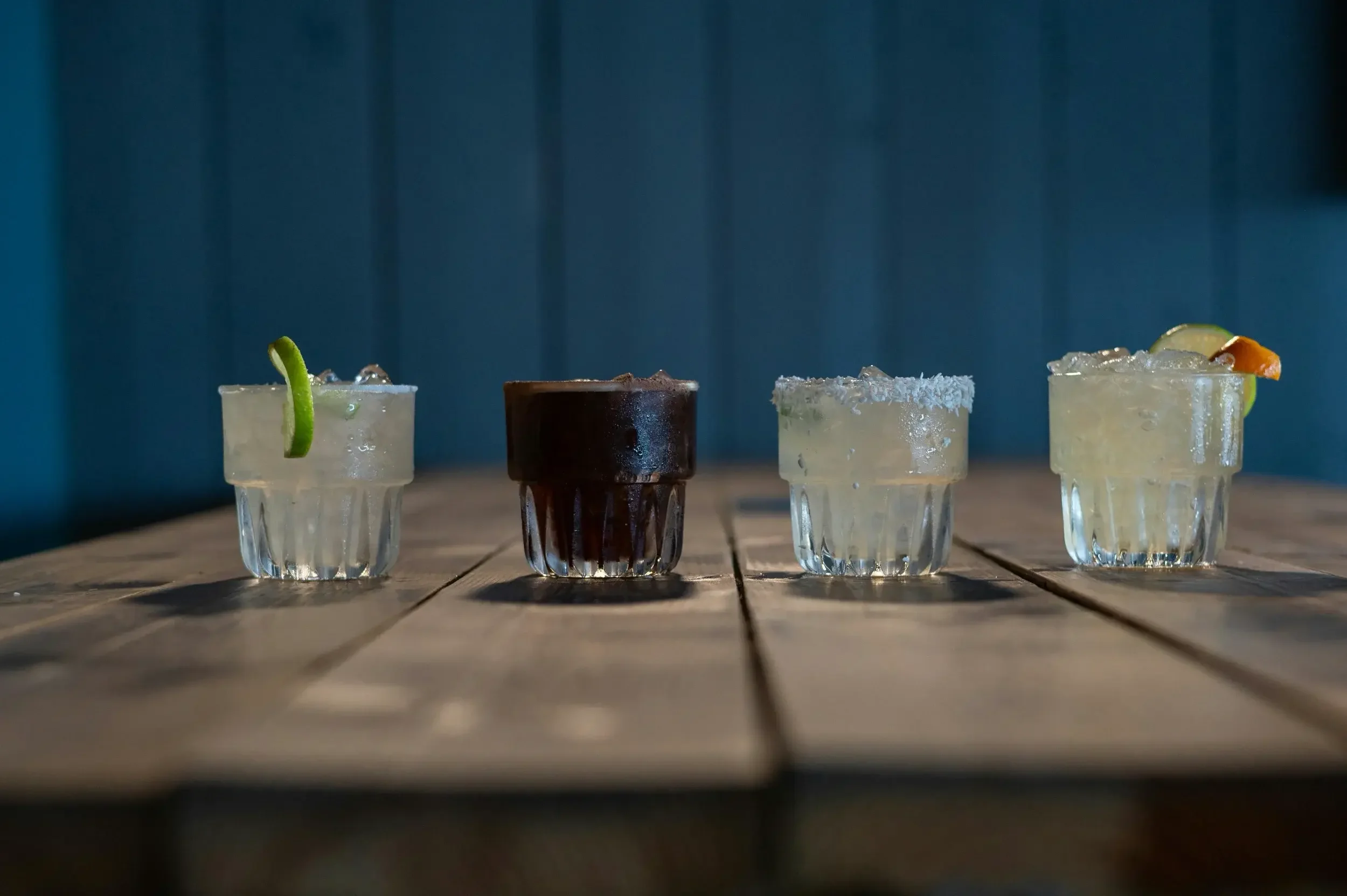 Four small glasses of cocktails on a wooden table against a blue wall. The first drink has a lime wedge, the second is dark, the third has a salted rim, and the fourth has a lime wedge and fruit garnish.