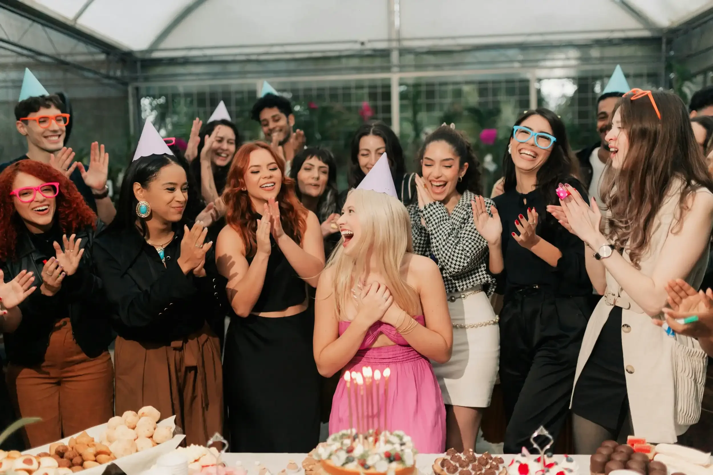 A group of people celebrating a birthday party indoors, with one woman in a pink dress front and center, smiling and surrounded by friends wearing colorful glasses and party hats, with a table of desserts in front.