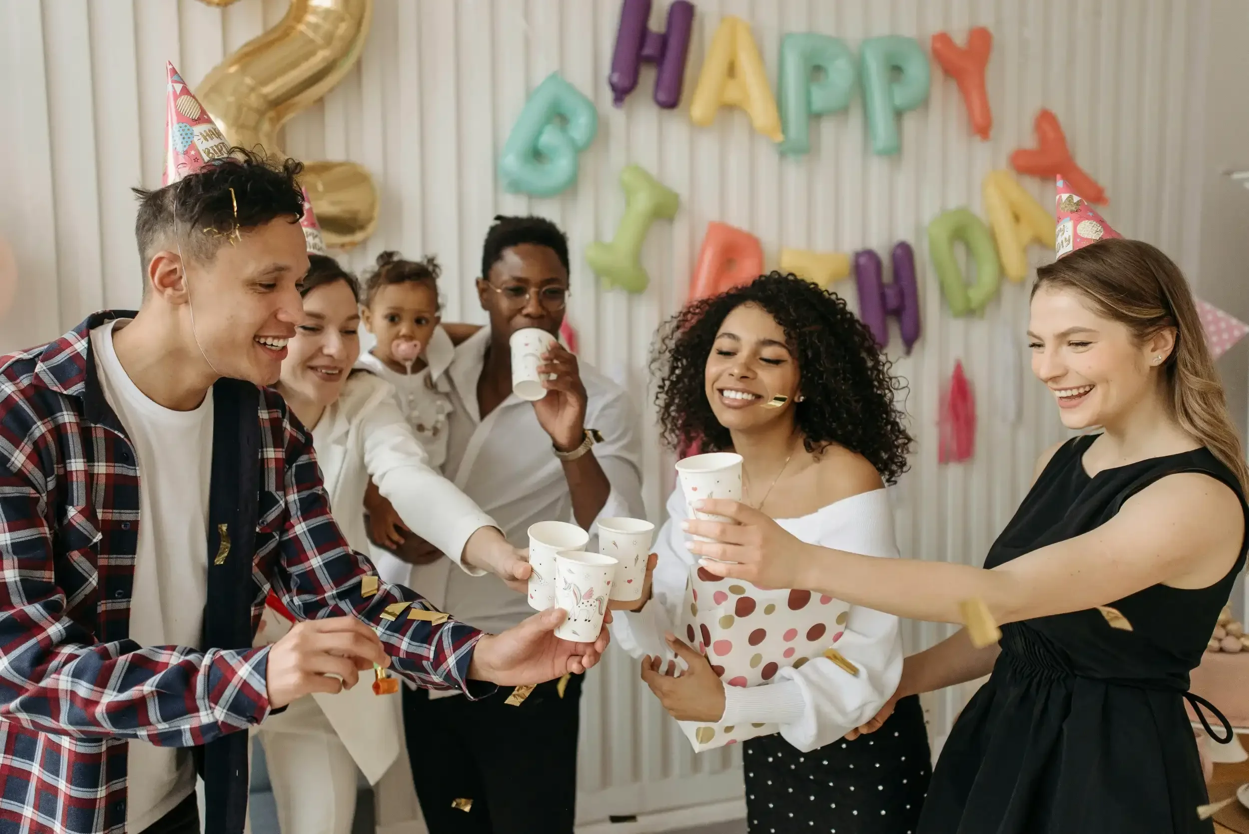 People celebrating a birthday party with colorful balloons and 'Happy Birthday' banner in the background, some wearing party hats, sharing drinks, and smiling.