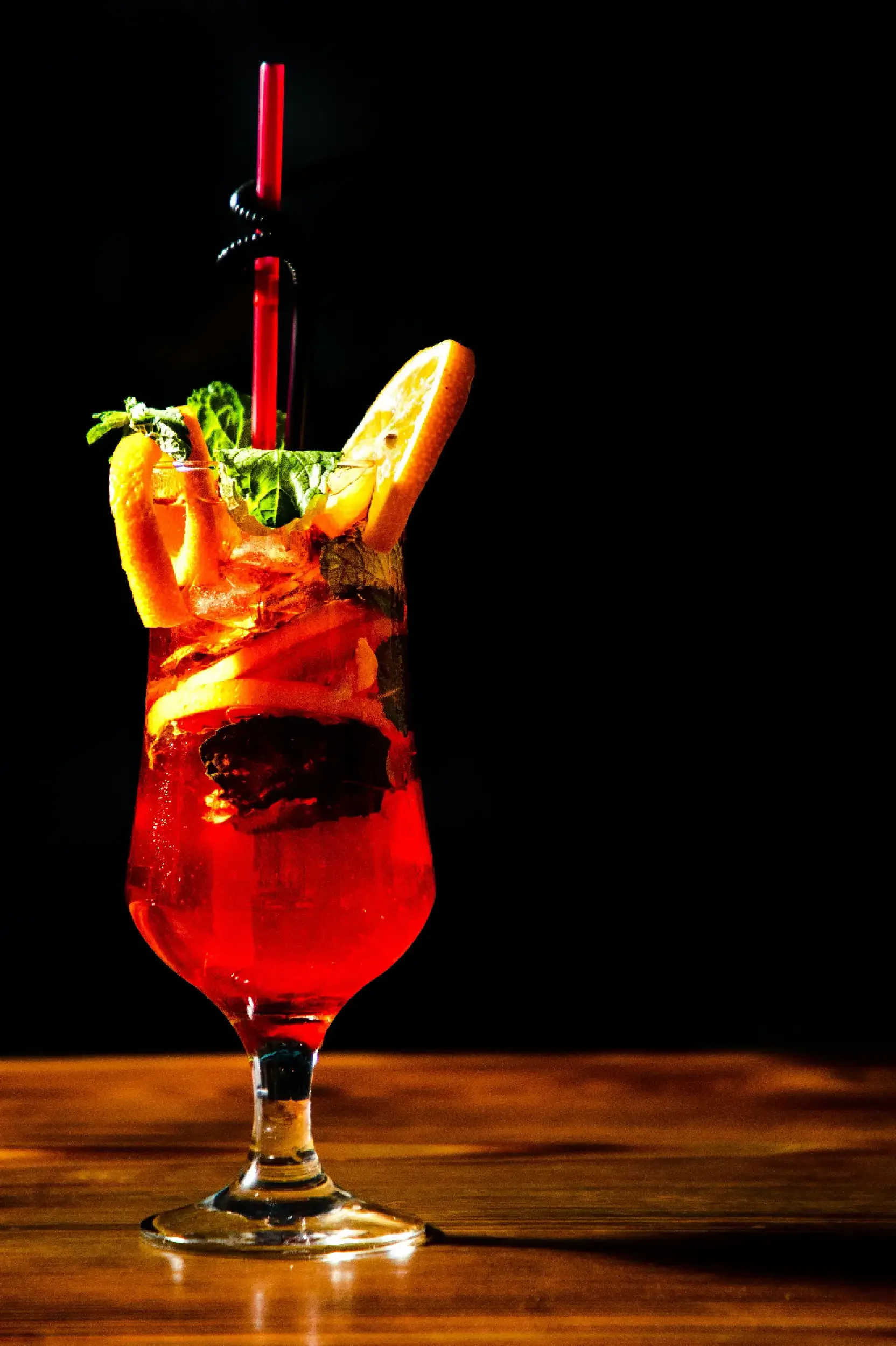 A tall glass with a red drink, garnished with lemon and orange slices, mint leaves, and a red straw, set on a wooden surface against a black background.