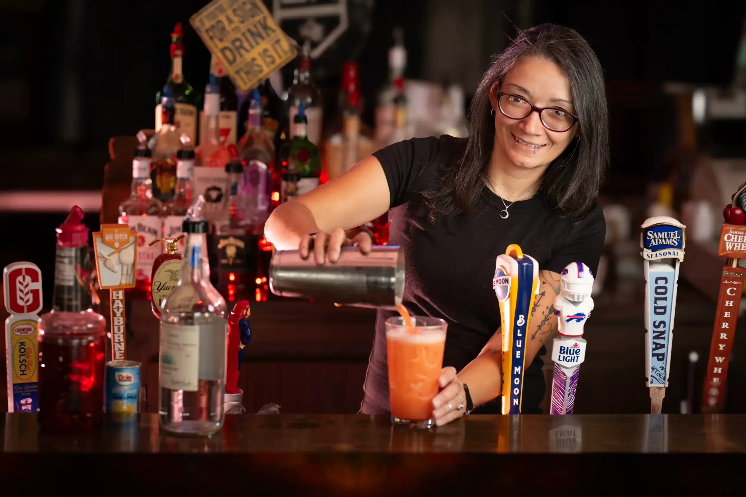 A woman with long dark hair and glasses making a cocktail at a bar, surrounded by various colorful beer tap handles and liquor bottles, smiling and looking to her right.
