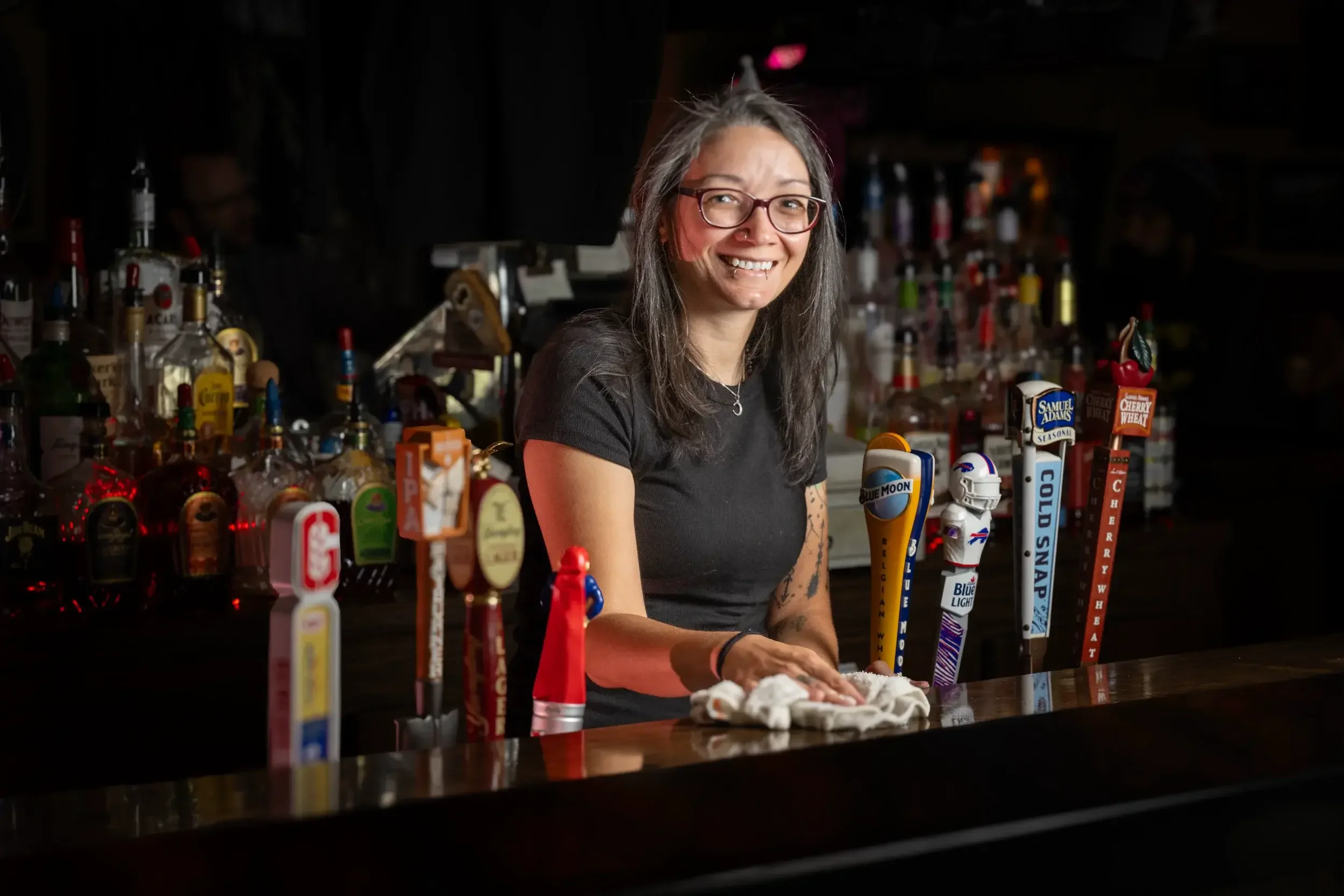Woman with long gray hair, glasses, and a nose ring smiling behind a bar counter with various beer taps.