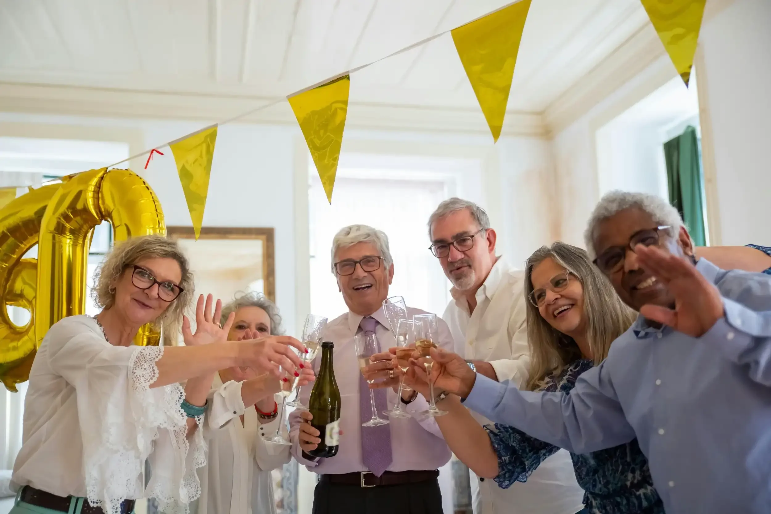 Group of elderly people celebrating at a party, raising glasses for a toast, with yellow banner flags and a large gold balloon in the background.