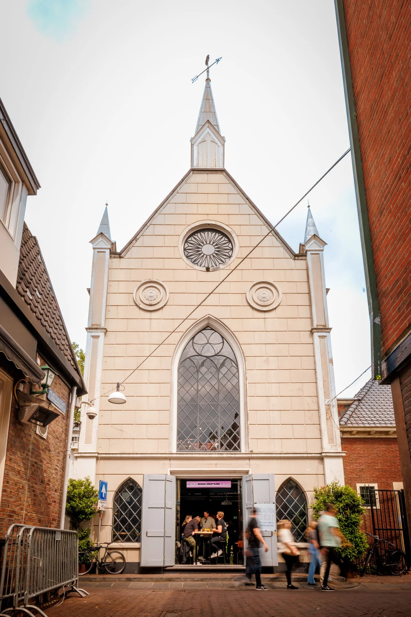 The facade of a church with a pointed roof and stained glass windows. People are standing and walking outside the church, some entering the building. The scene appears lively with a group gathered just outside the entrance.