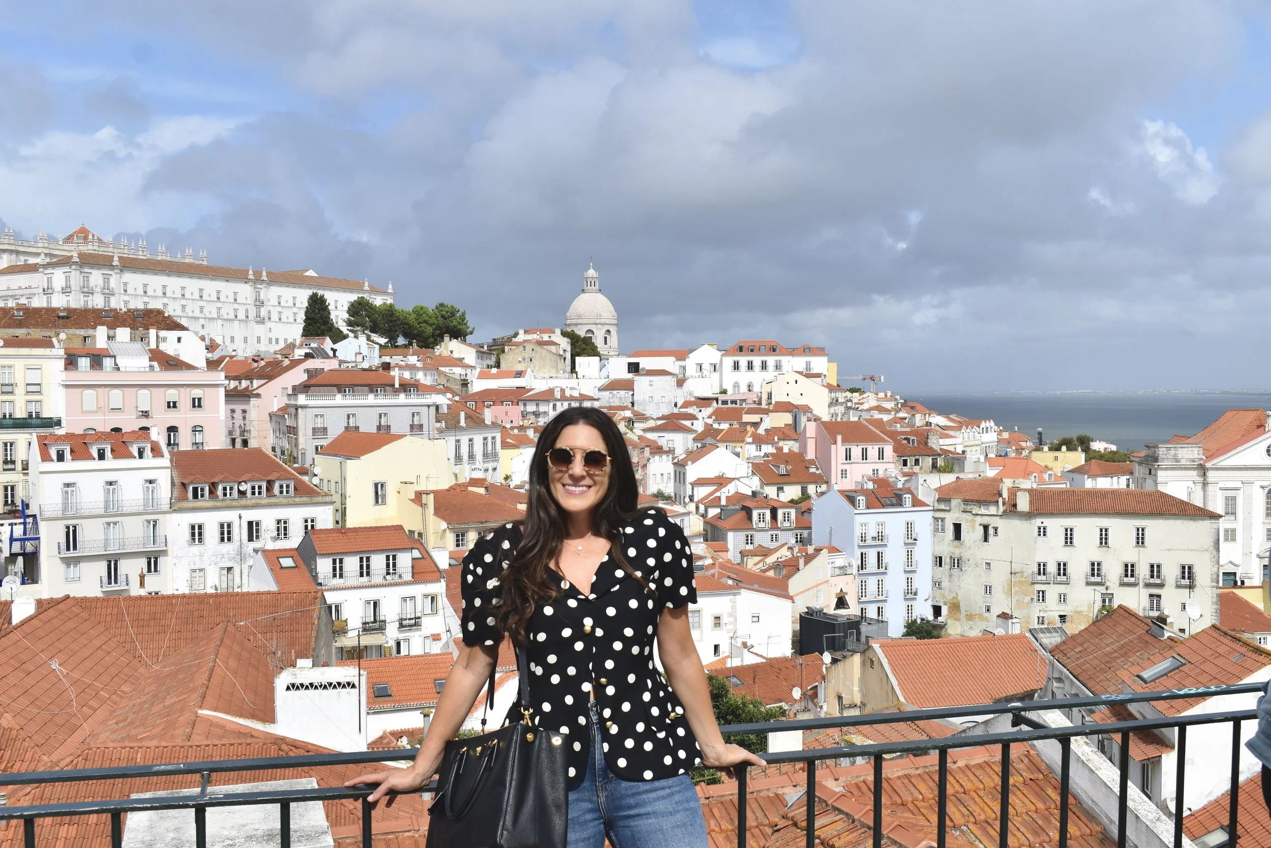 A woman with long dark hair wearing sunglasses and a black polka dot top stands on a balcony overlooking a cityscape with reddish rooftops, a prominent white domed building, and the sea in the background under a partly cloudy sky.
