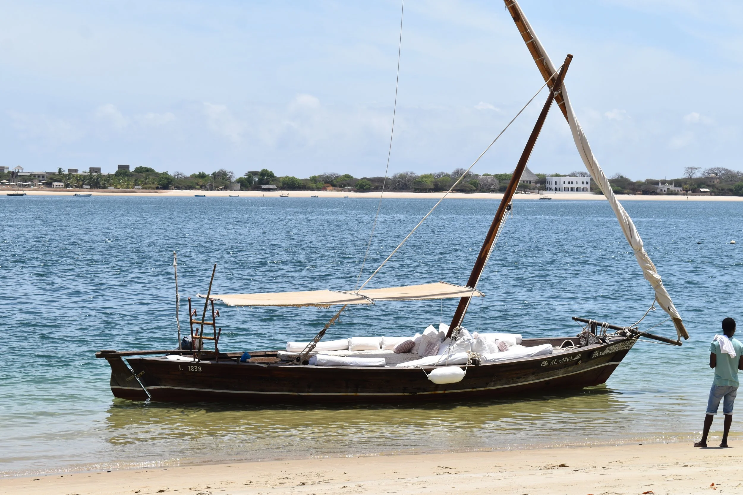 A traditional wooden sailboat with a single mast near the shore, with some people standing on the beach and distant buildings and trees across the water.
