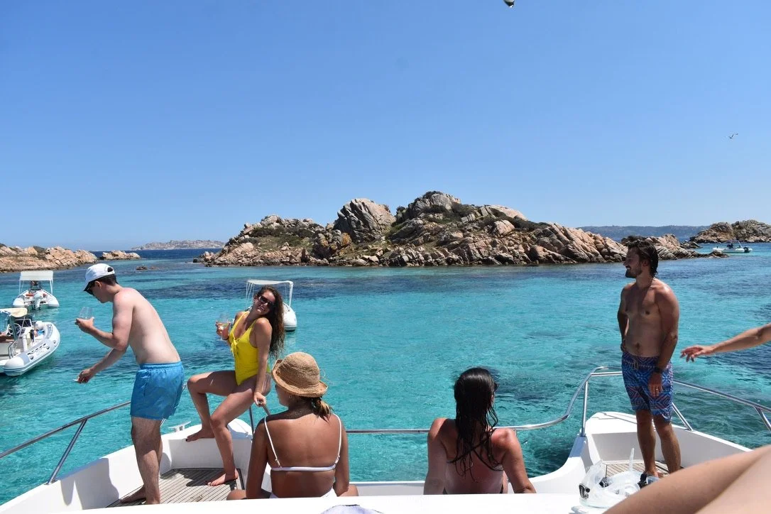 Group of people on a boat floating on turquoise water near rocky islands, some standing and others sitting, under a clear blue sky.