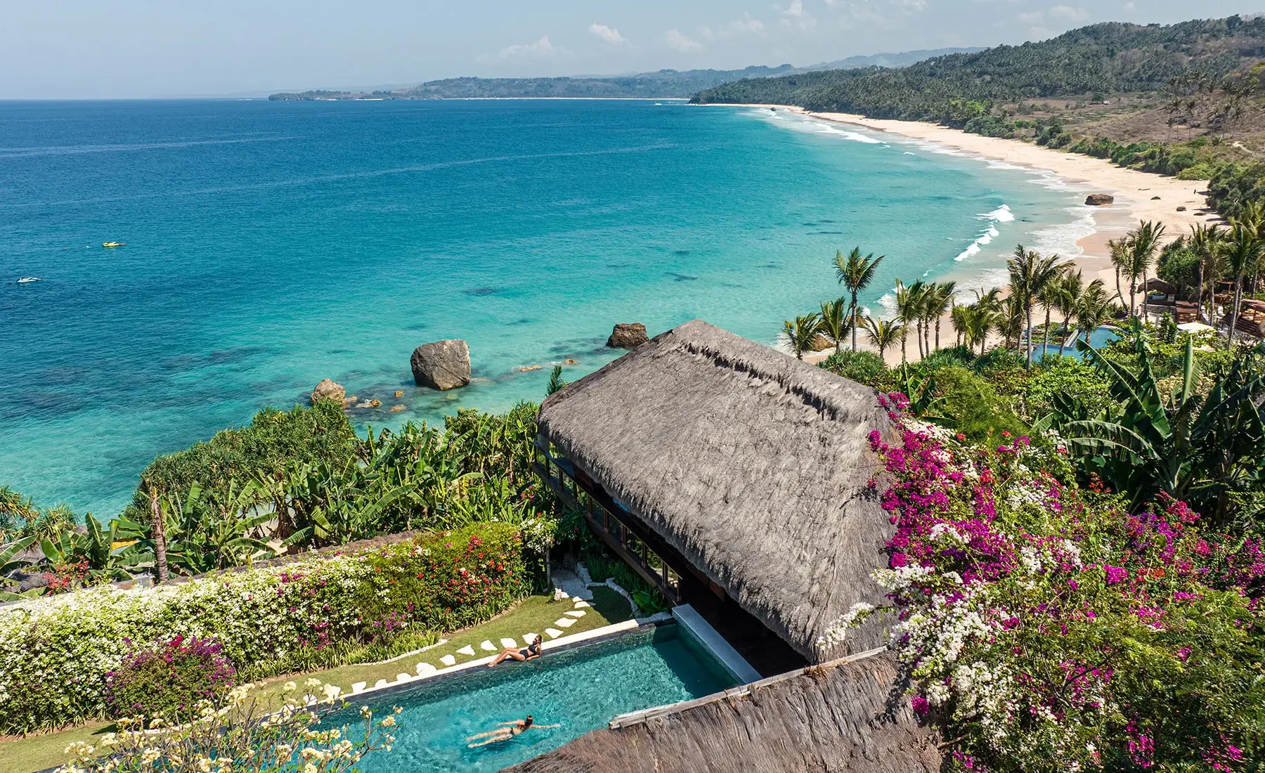 A tropical beach with turquoise water and white sand, lined with palm trees and lush greenery. A thatched-roof building over a pool is in the foreground, with colorful flowers and two people relaxing near the pool. Large rocks are in the water near the shore, and distant hills extend into the ocean under a partly cloudy sky.