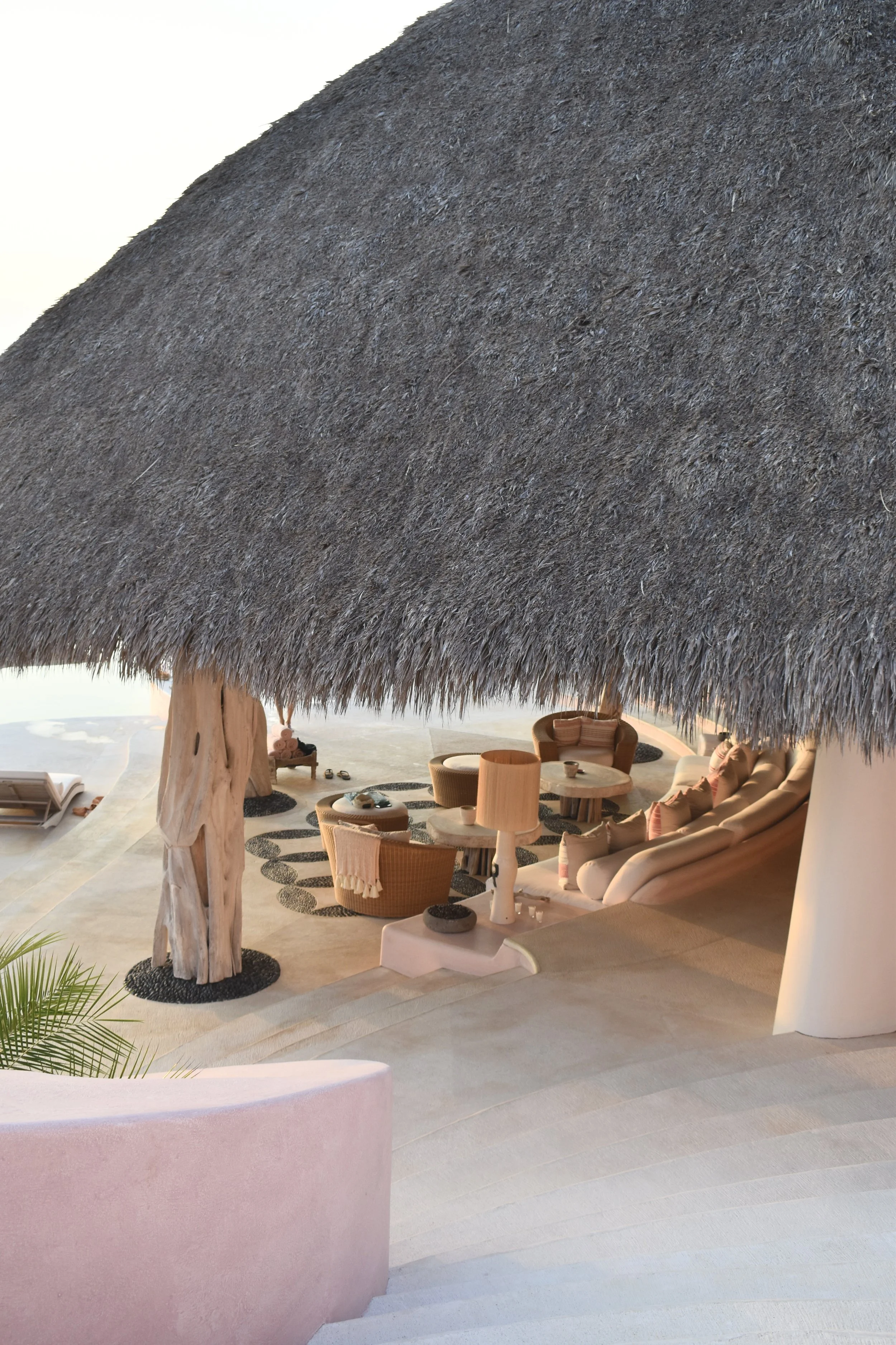 A thatched roof over a lounge area with wicker chairs, white cushions, and small tables on a sandy floor, with a pink planter and some greenery in the foreground.