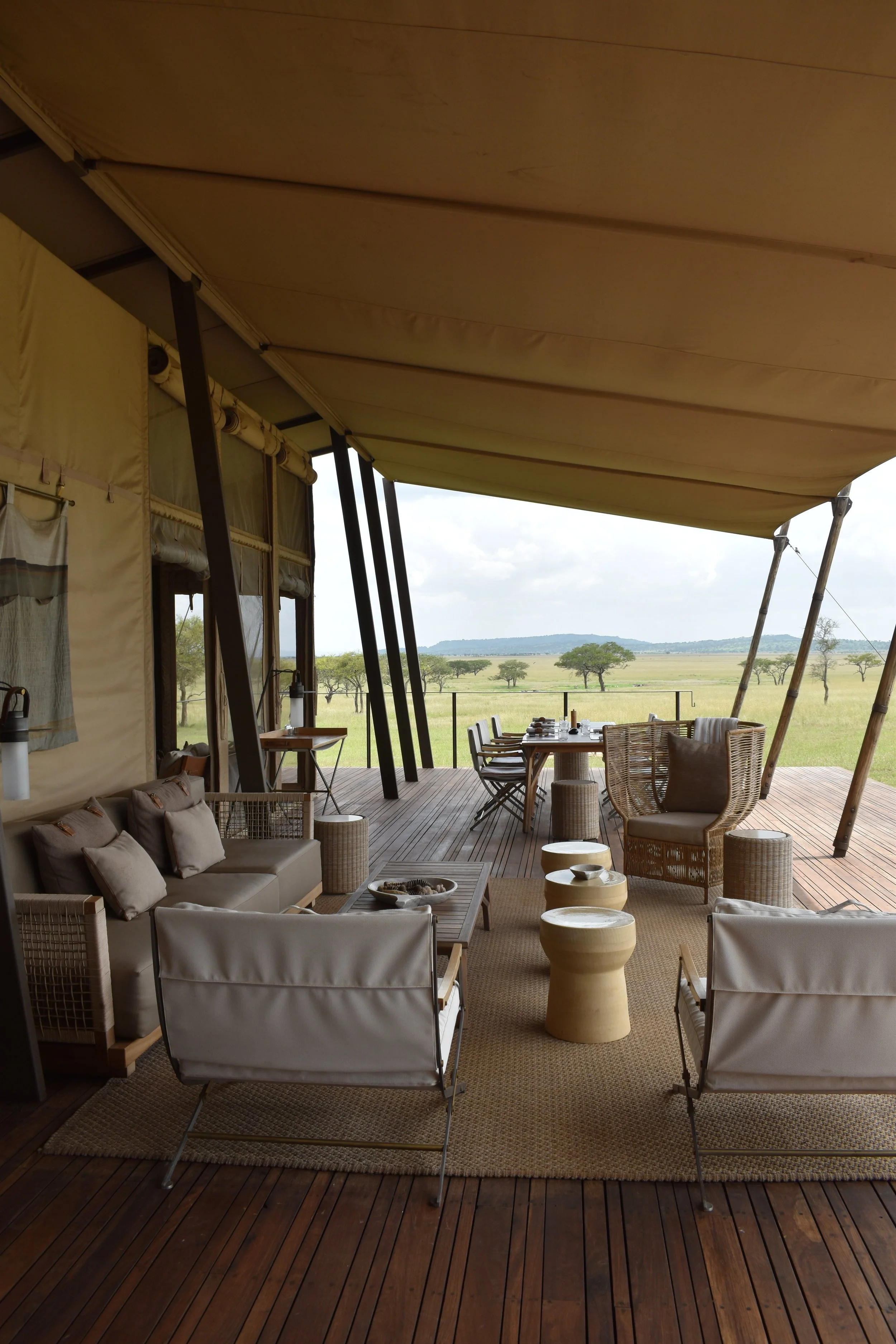 A covered outdoor patio area with modern furniture, including chairs, sofas, and round tables, overlooking a vast open savannah landscape with scattered trees under a cloudy sky.