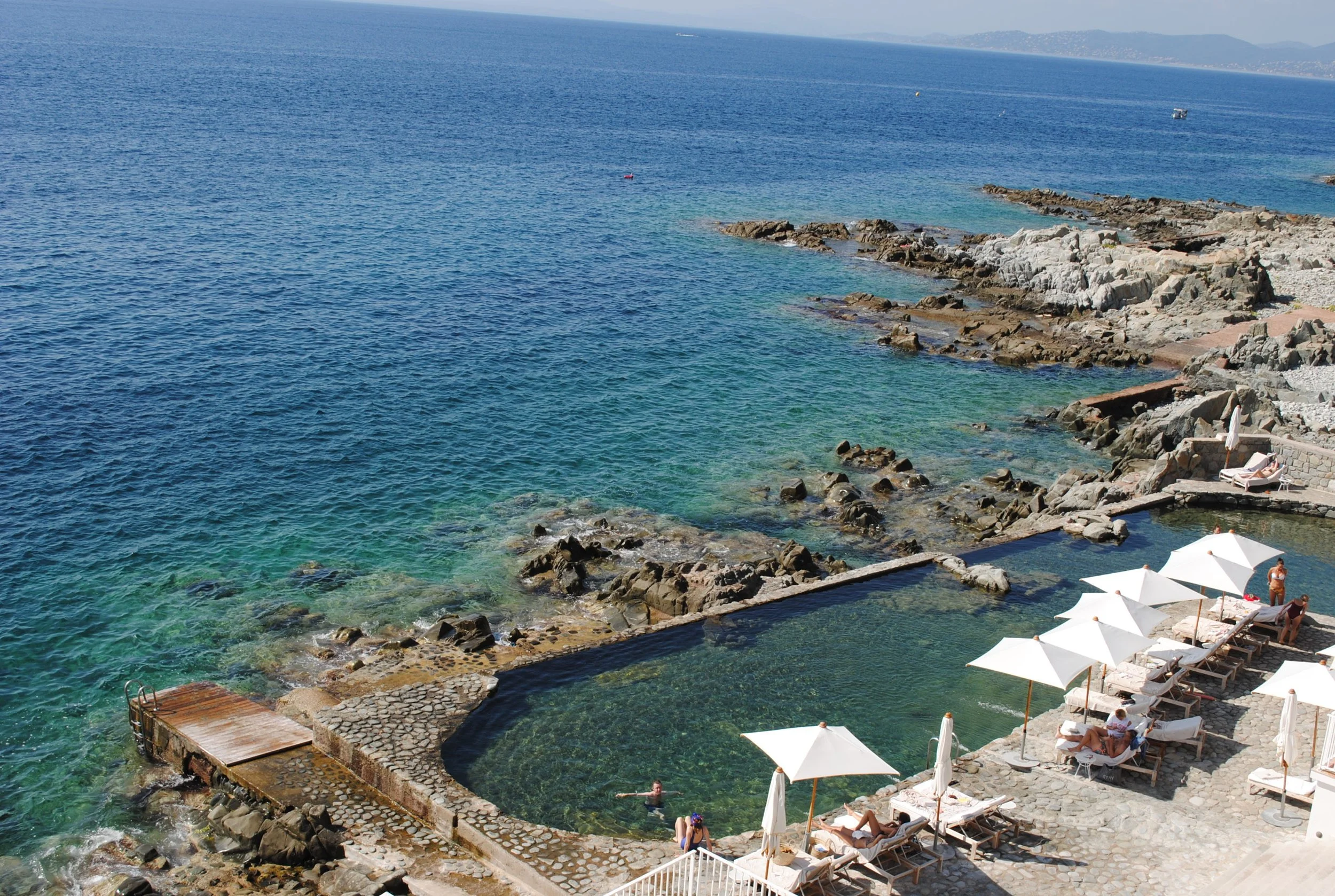 A coastal scene with clear blue water, rocky shoreline, and white umbrellas with lounge chairs, where people are sunbathing and swimming.