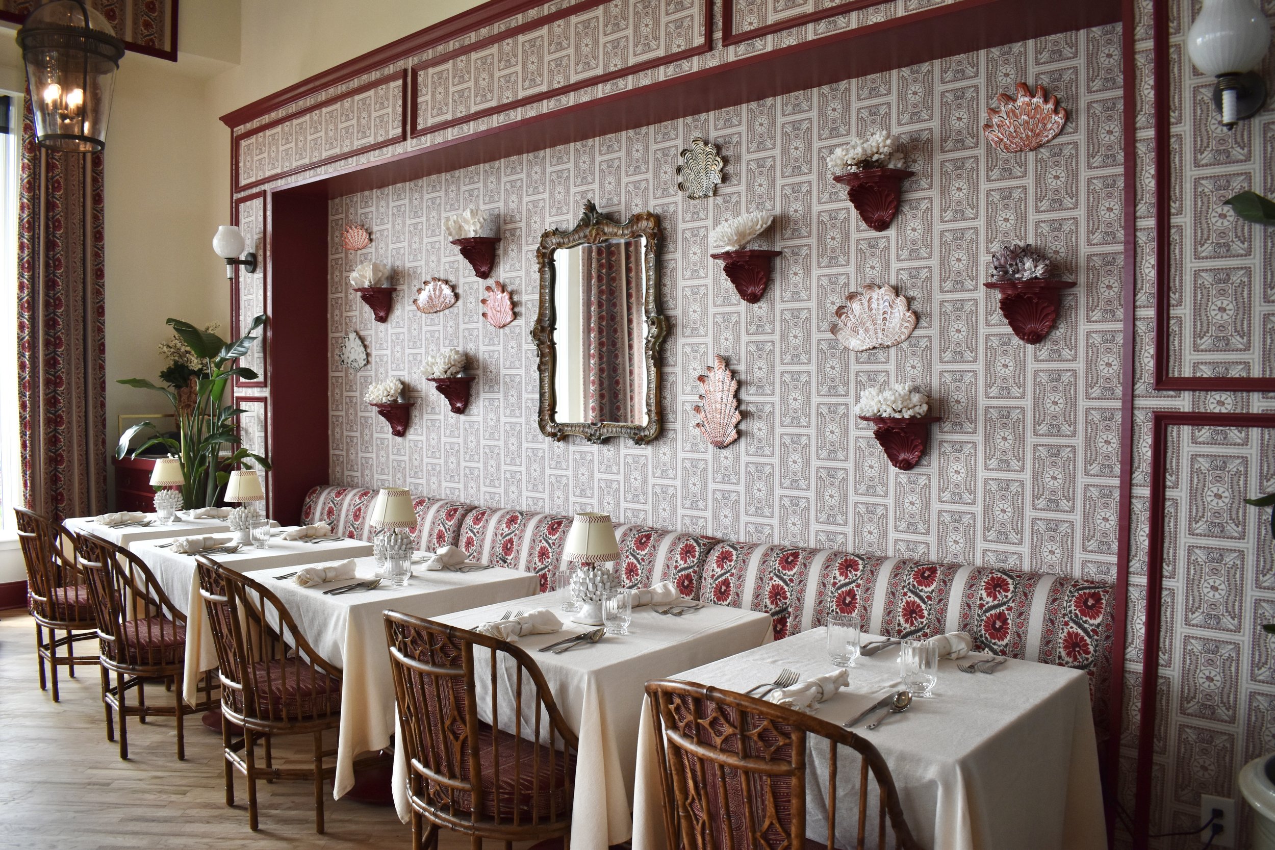 A dining room with a long table set with white tablecloths, glasses, silverware, and napkins. The chairs are wooden with red cushions. The wall behind features ornate wallpaper and shelves holding decorative shells and a mirror.