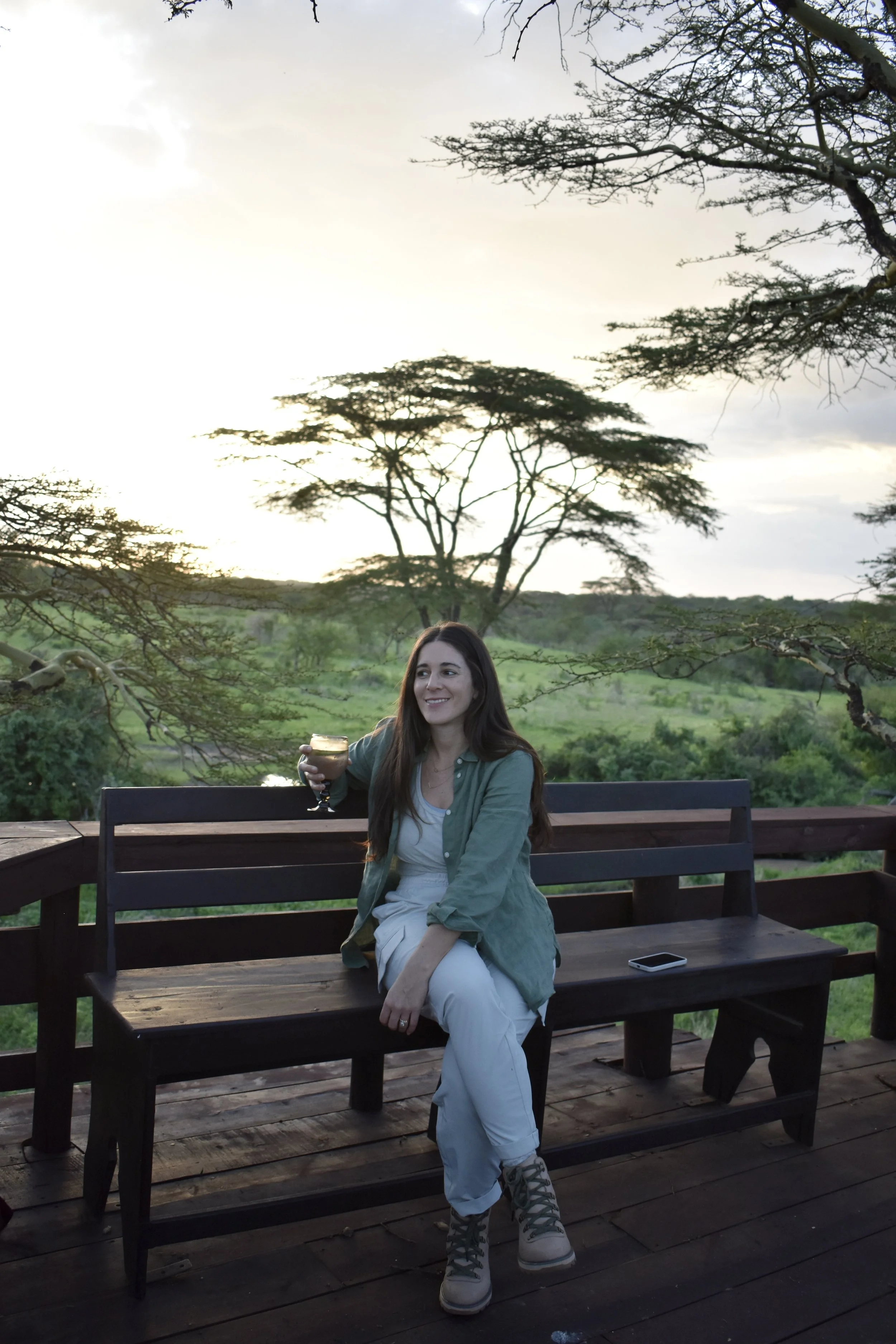 A woman with long dark hair sitting on a wooden park bench outdoors, holding a drink in her right hand, smiling, wearing a green shirt over a white top, white pants, and grey hiking boots, with a cellphone on the bench beside her. The background shows green trees and a cloudy sky at sunset.