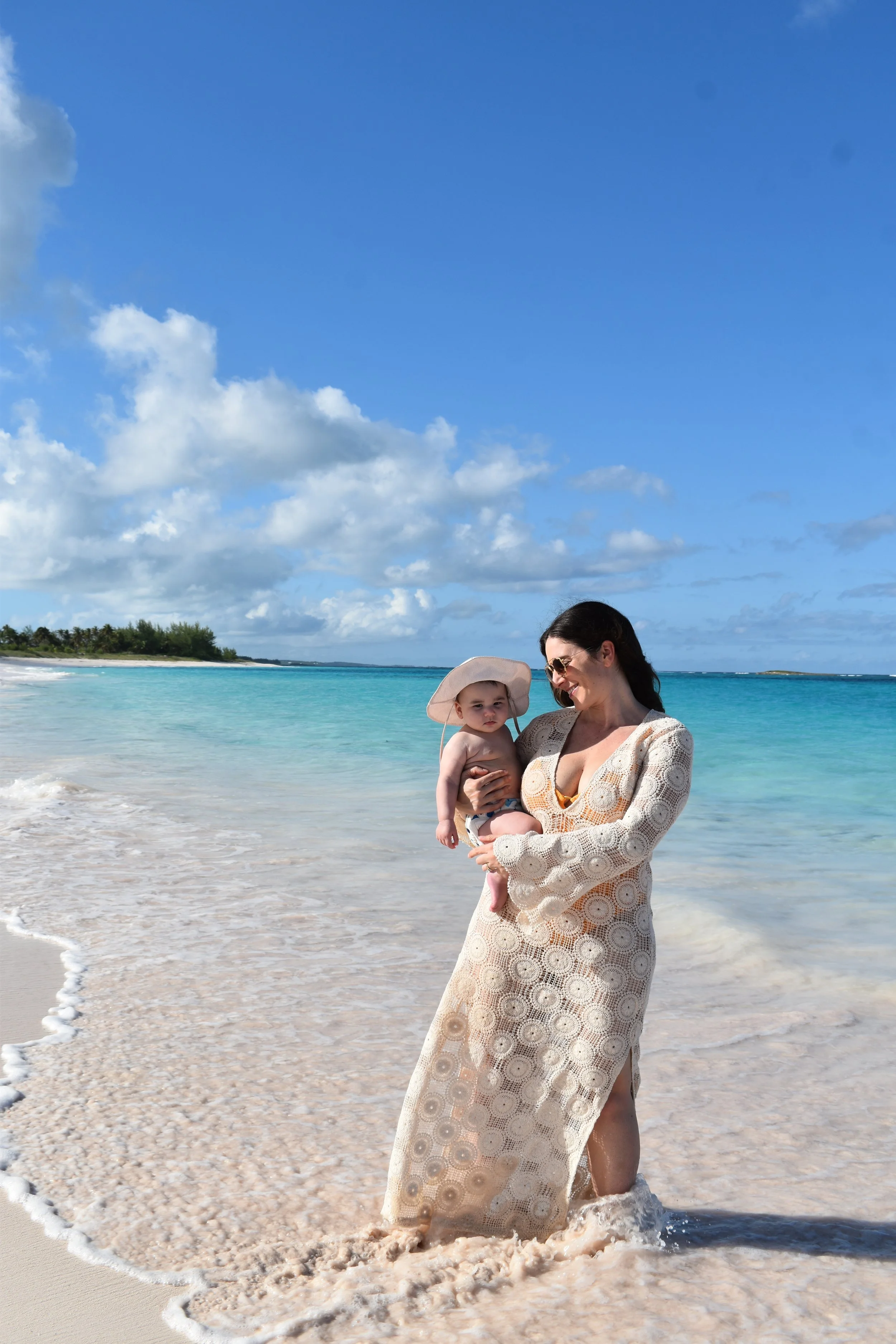 Woman holding a young child at the beach, both dressed in light clothing suitable for warm weather, with the ocean, sandy shore, and a partly cloudy sky in the background.