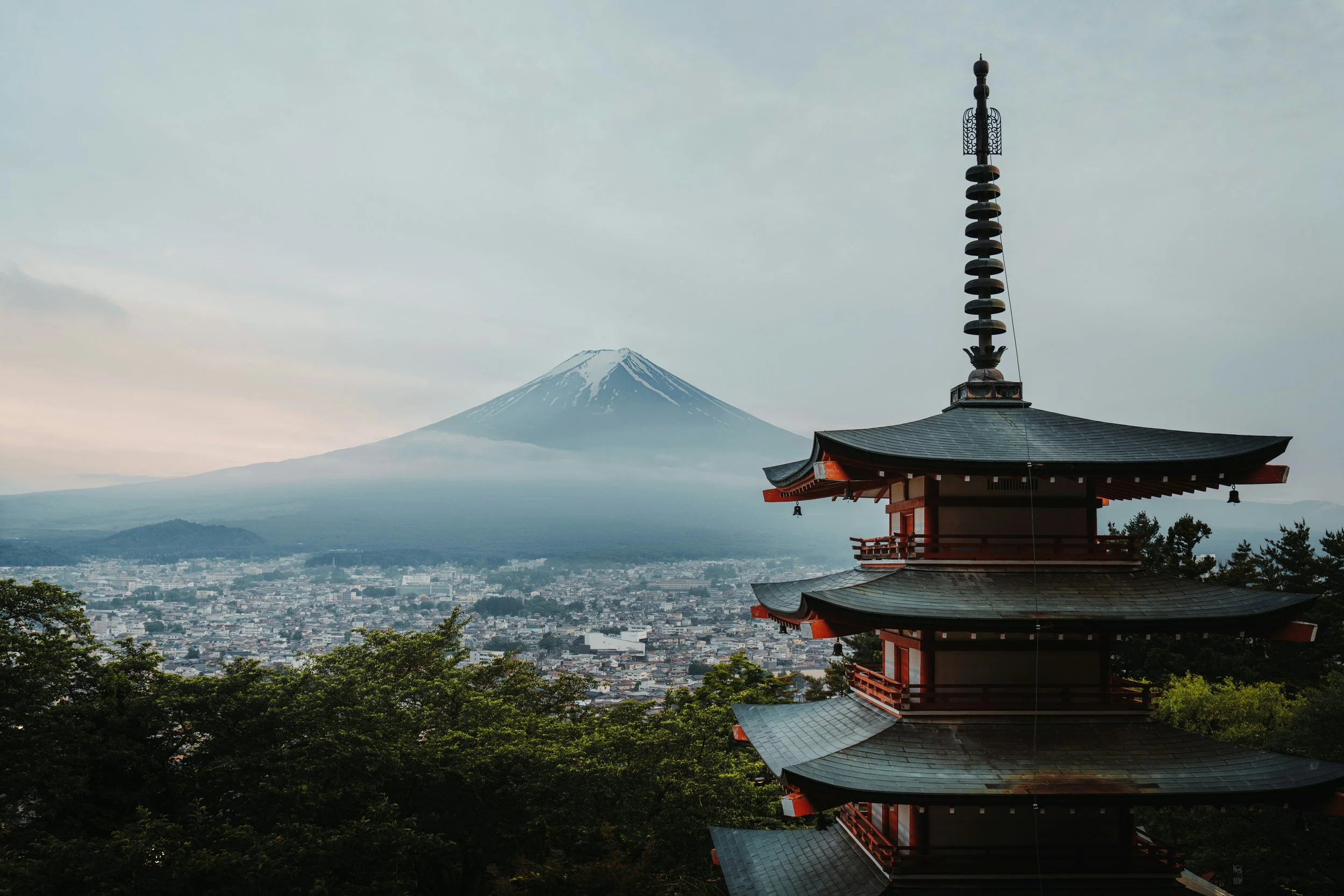 A pagoda-style building with a tall, ornate spire in the foreground, Mount Fuji with snow on its peak in the background, and a cityscape below.