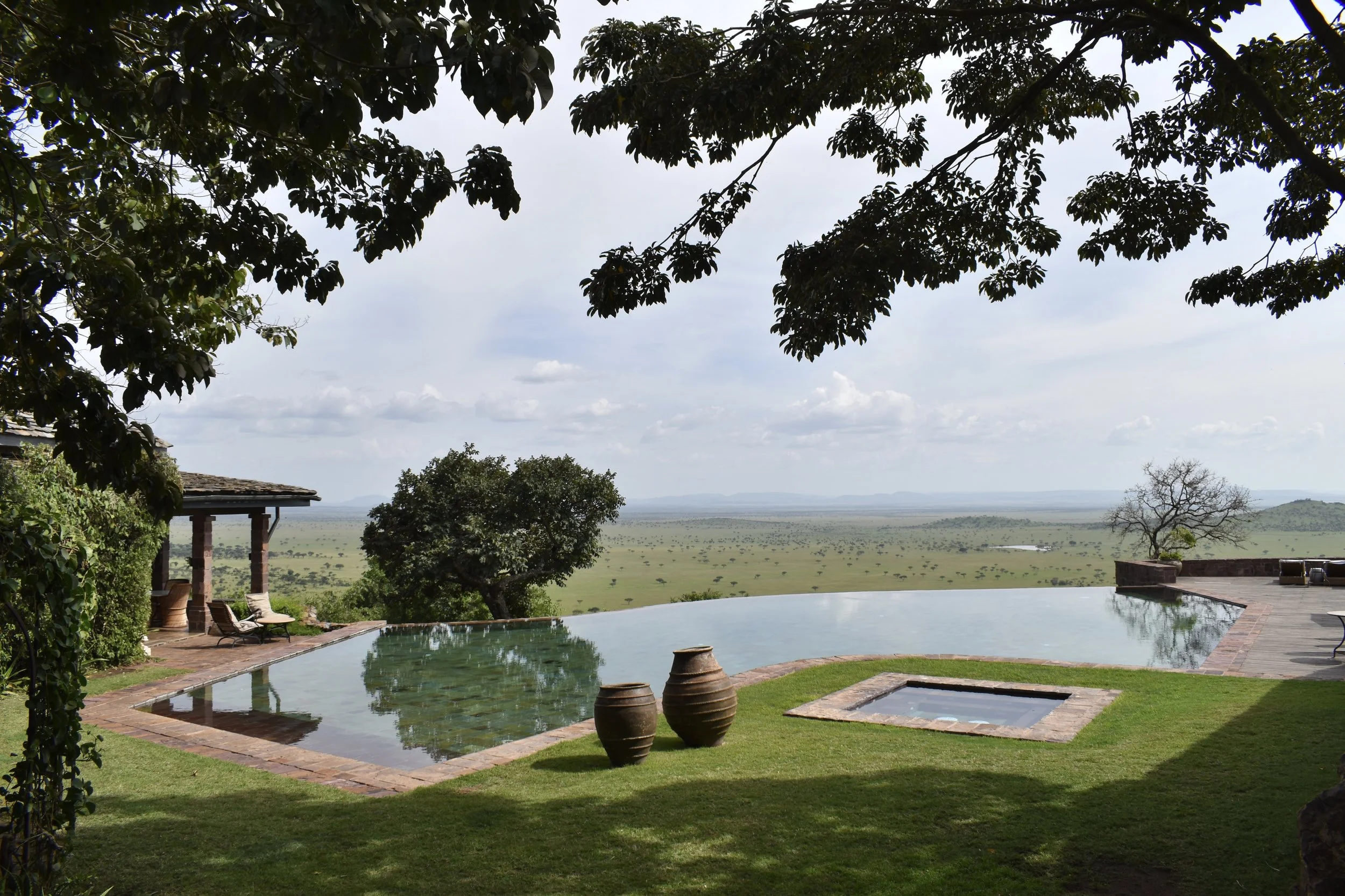 View of an outdoor pool area with lounge chairs, large clay pots, and a scenic landscape with trees and open plains in the distance under cloudy skies.