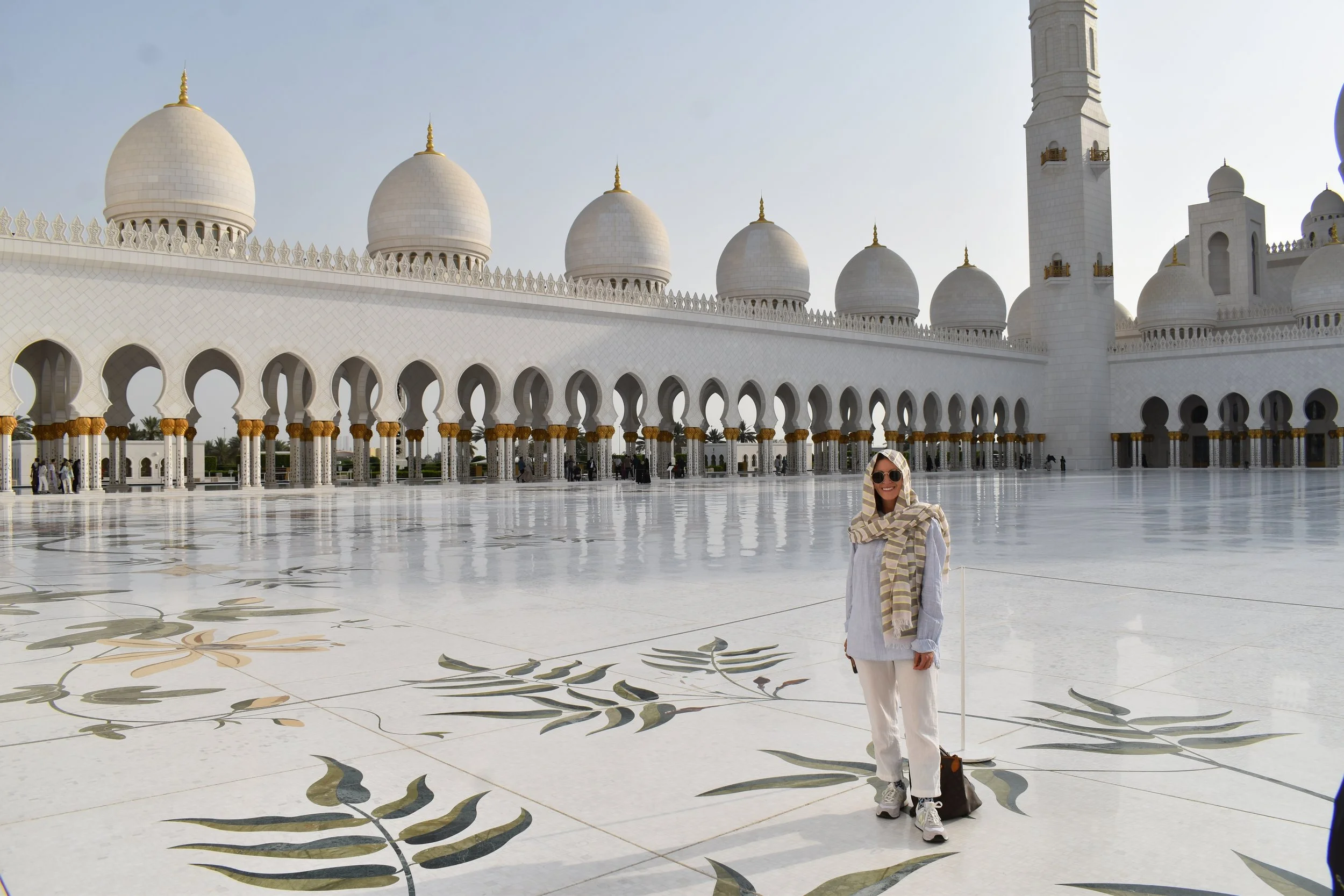 A woman in white clothing and sunglasses standing in front of the Sheikh Zayed Grand Mosque in Abu Dhabi, with a large marble courtyard decorated with floral patterns and multiple domes and minarets in the background.