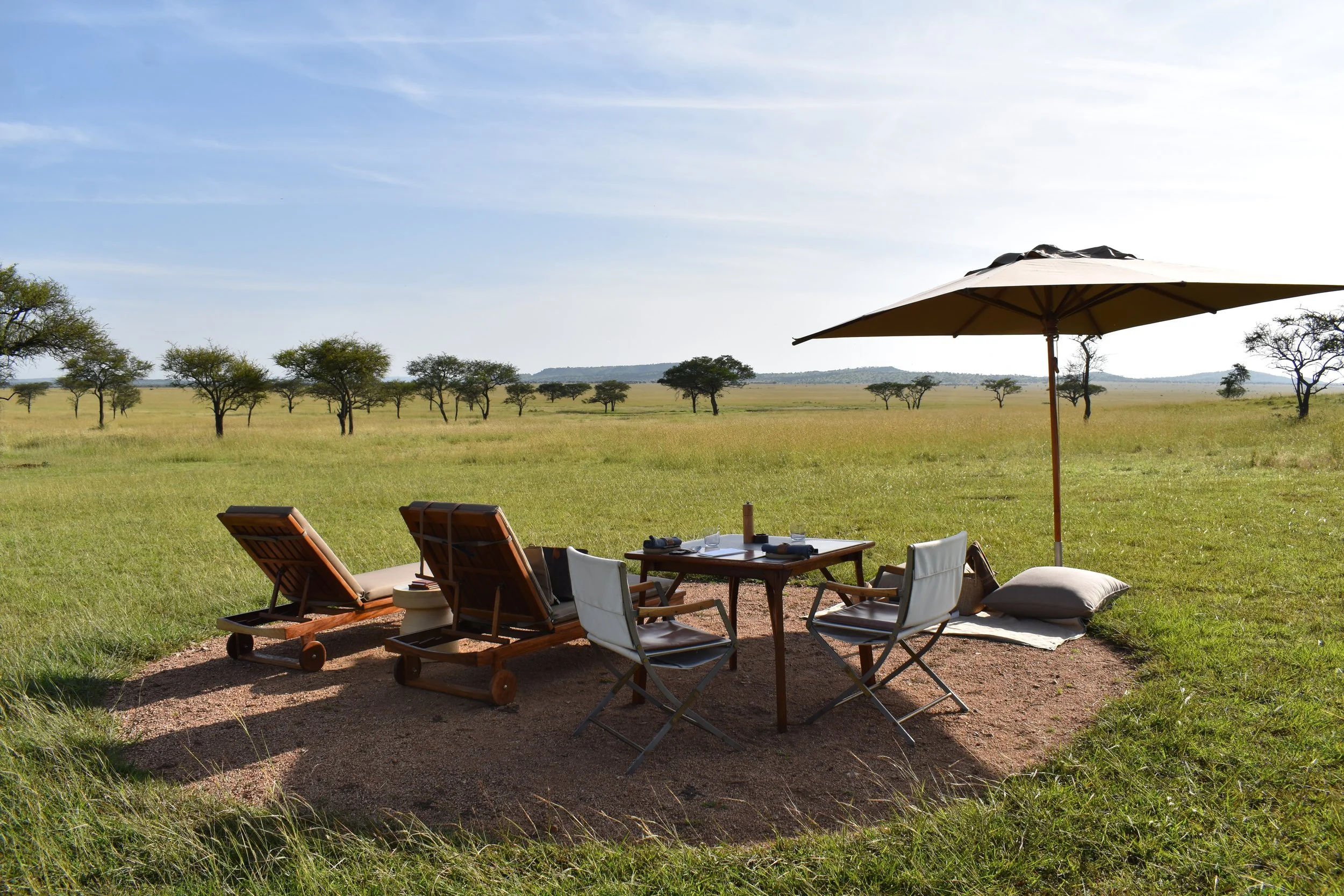 Outdoor dining setup in a safari landscape with a large beige umbrella, chairs, a table set for meals, and distant trees under a clear blue sky.