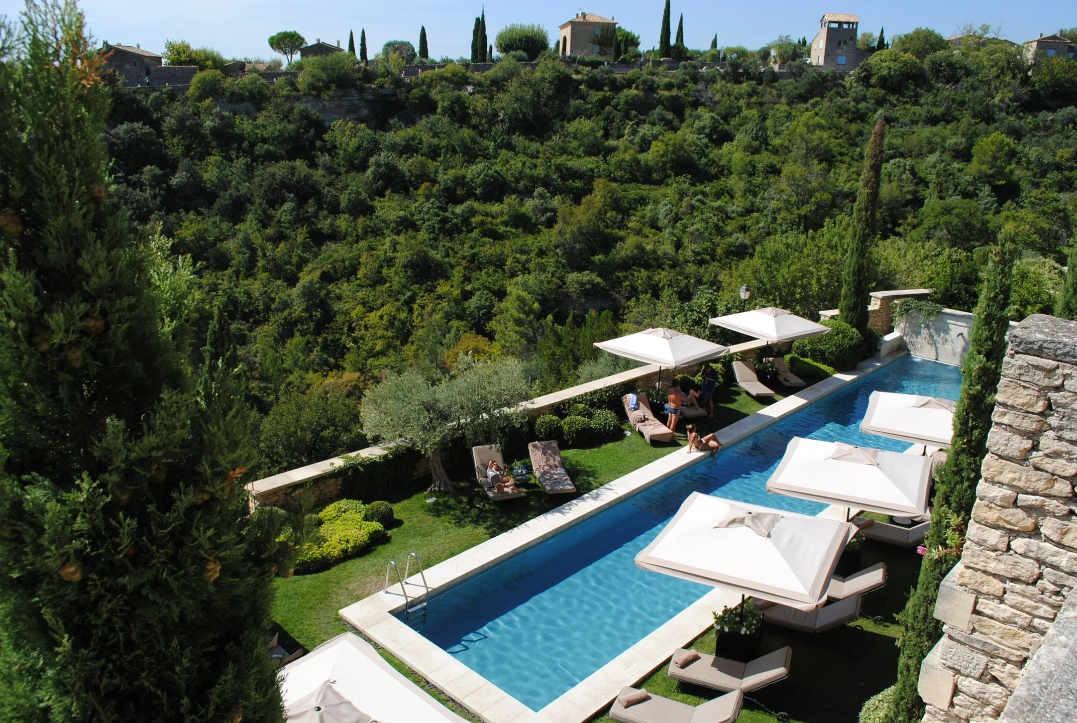 Luxury poolside area with lounge chairs and umbrellas overlooking a lush green hillside with houses and cypress trees.