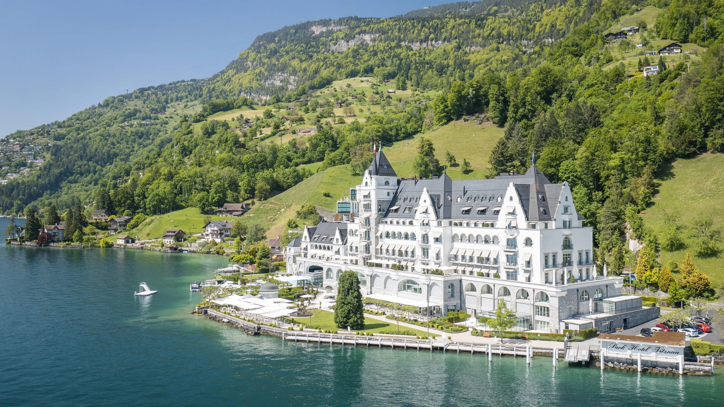 A large white hotel building with a steep black roof sits at the edge of a lake, surrounded by lush green hills with houses scattered across them. The hotel features multiple towers and balconies, and the shoreline has a walking path and outdoor seating areas.