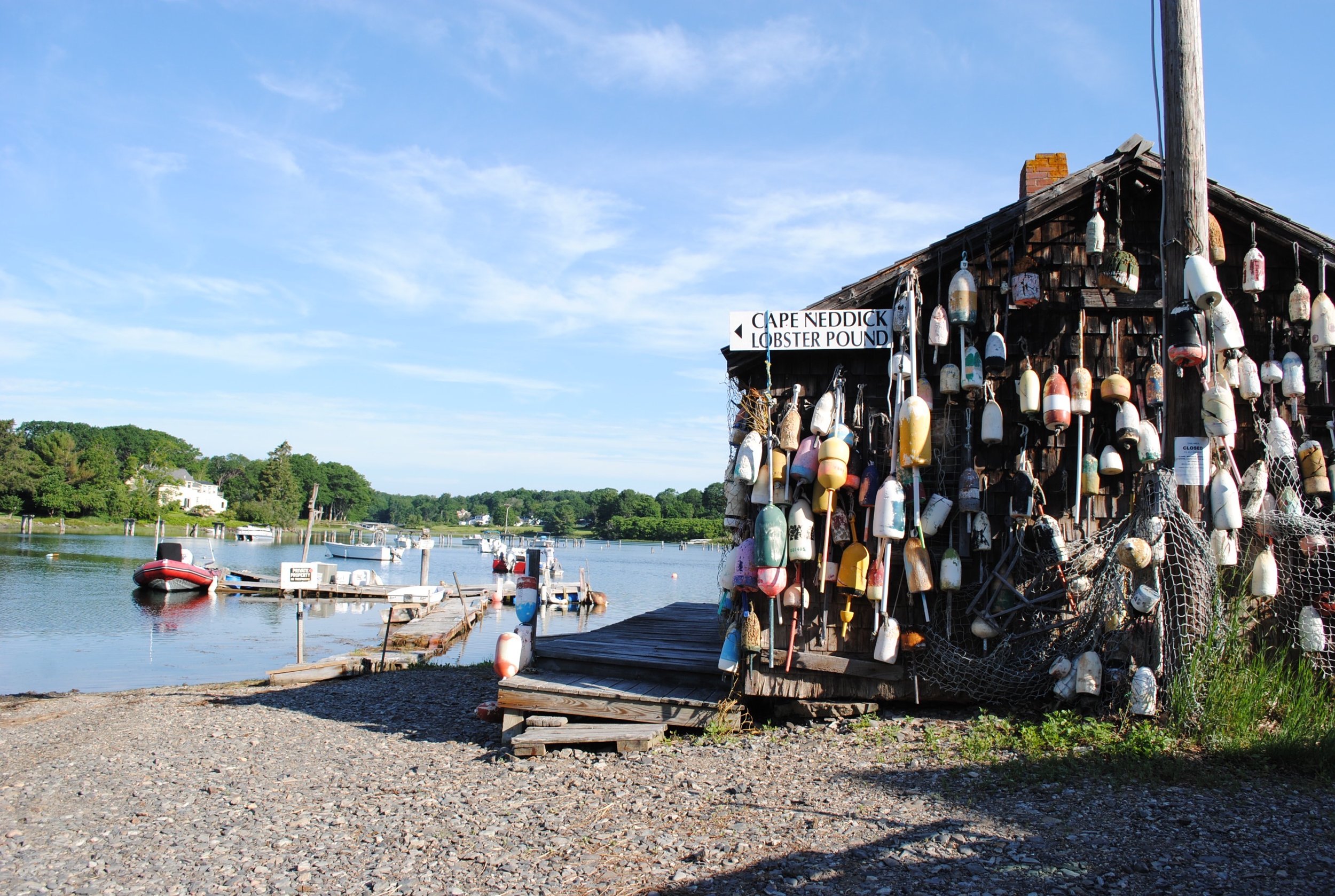 A wooden structure decorated with many colorful buoys hangs off the side of a dock near a calm body of water. In the background, boats are floating on the water with trees and houses along the shoreline under a blue sky.