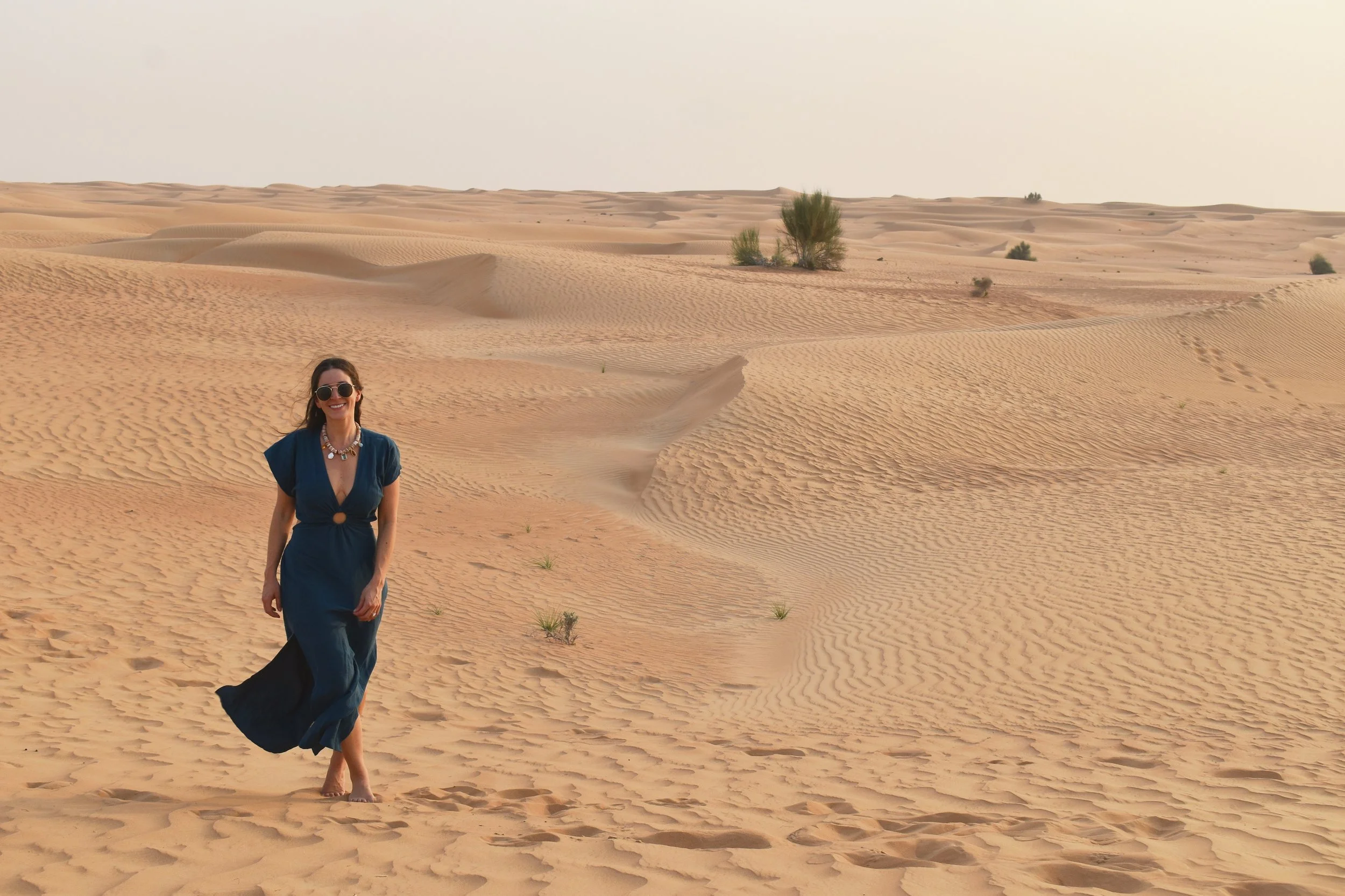 A woman in a long blue dress walking barefoot in a desert with sand dunes and sparse green bushes, wearing sunglasses and a necklace, smiling.