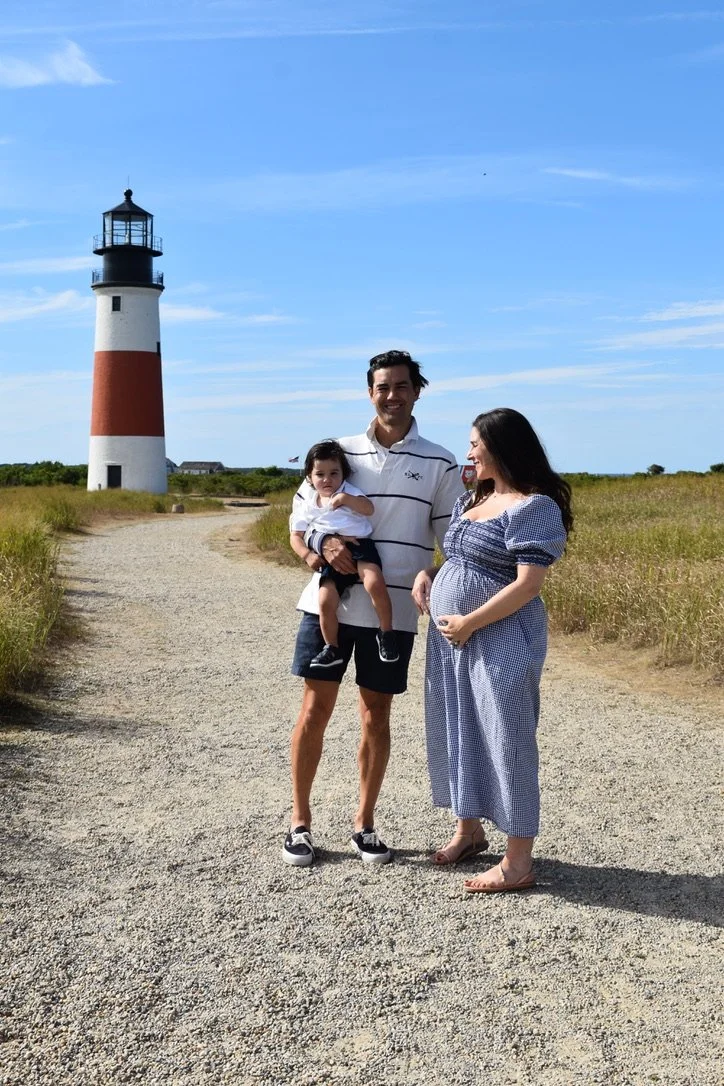 A family of three, including a pregnant woman, a man holding a child, standing on a gravel path near a lighthouse under a blue sky.