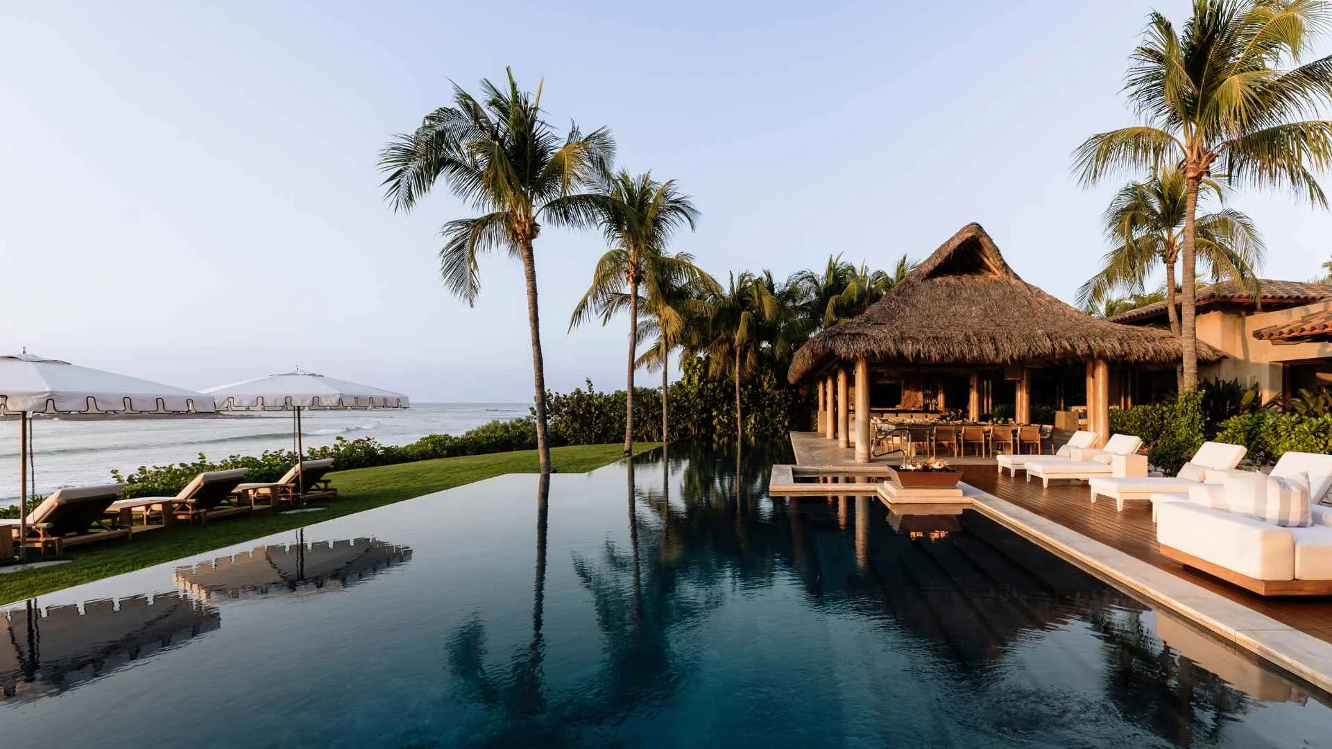 Luxury beachfront resort with a pool, palm trees, thatched-roof bar, lounge chairs, and ocean views at sunset.