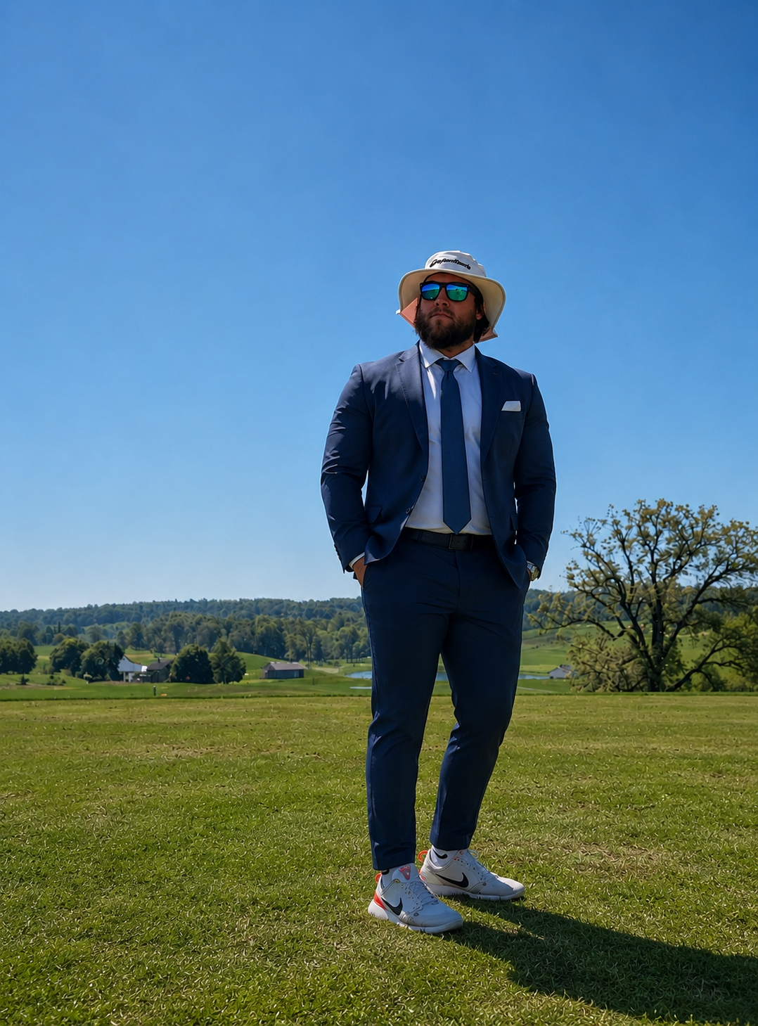 A man in a navy suit, tie, and sunglasses standing outdoors on a grassy field under a clear blue sky, with rolling hills and trees in the background.