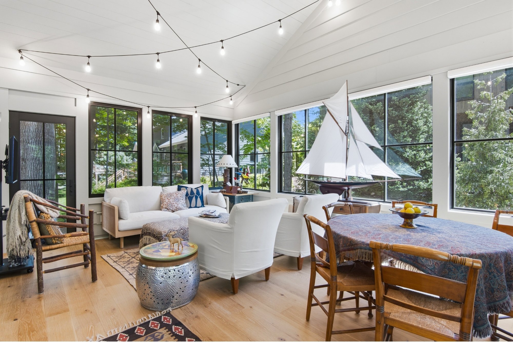 Sunroom with large windows, white ceiling with string lights, white sofas, rocking chairs, a sailing ship model, a dining table with a tablecloth, and various decorative items including a bowl of lemons.