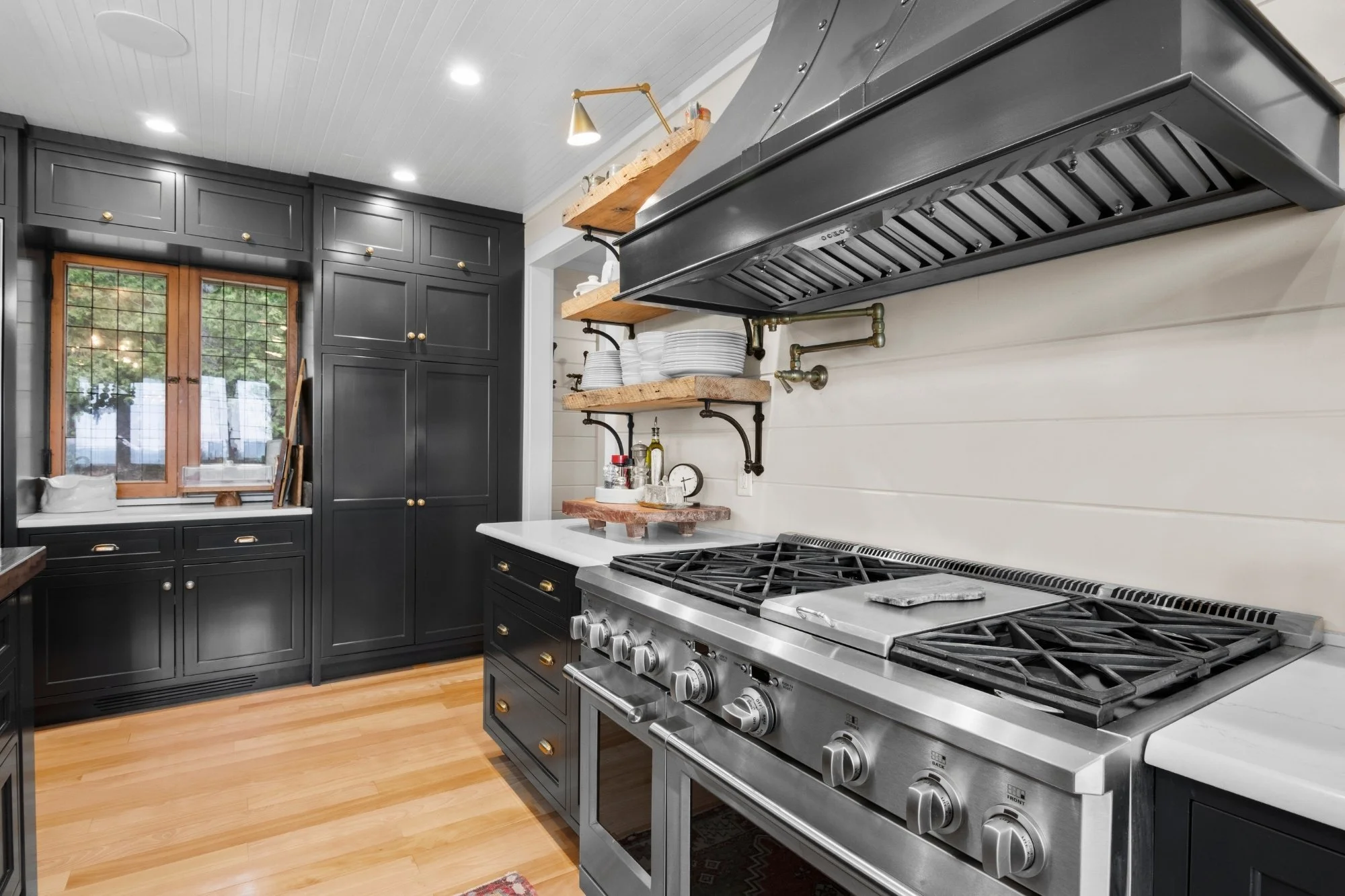 Modern kitchen with black cabinetry, wooden shelves holding dishes, a stainless steel gas stove, and a window with wooden frame, hardwood flooring, and a white ceiling with recessed lighting.