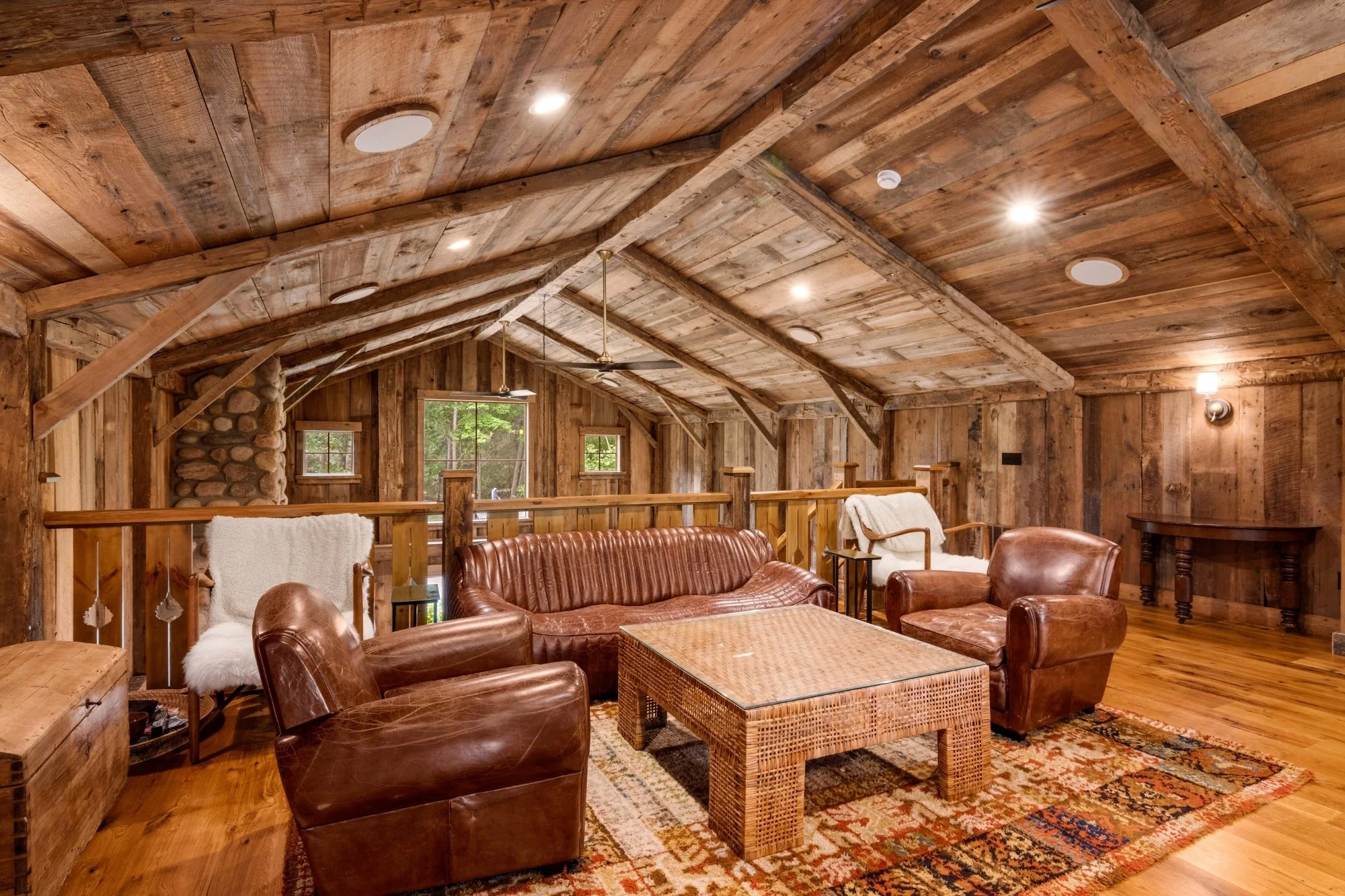Living room with wooden walls and ceiling, leather chairs, a wooden coffee table, patterned rug, and large windows showing greenery outside.