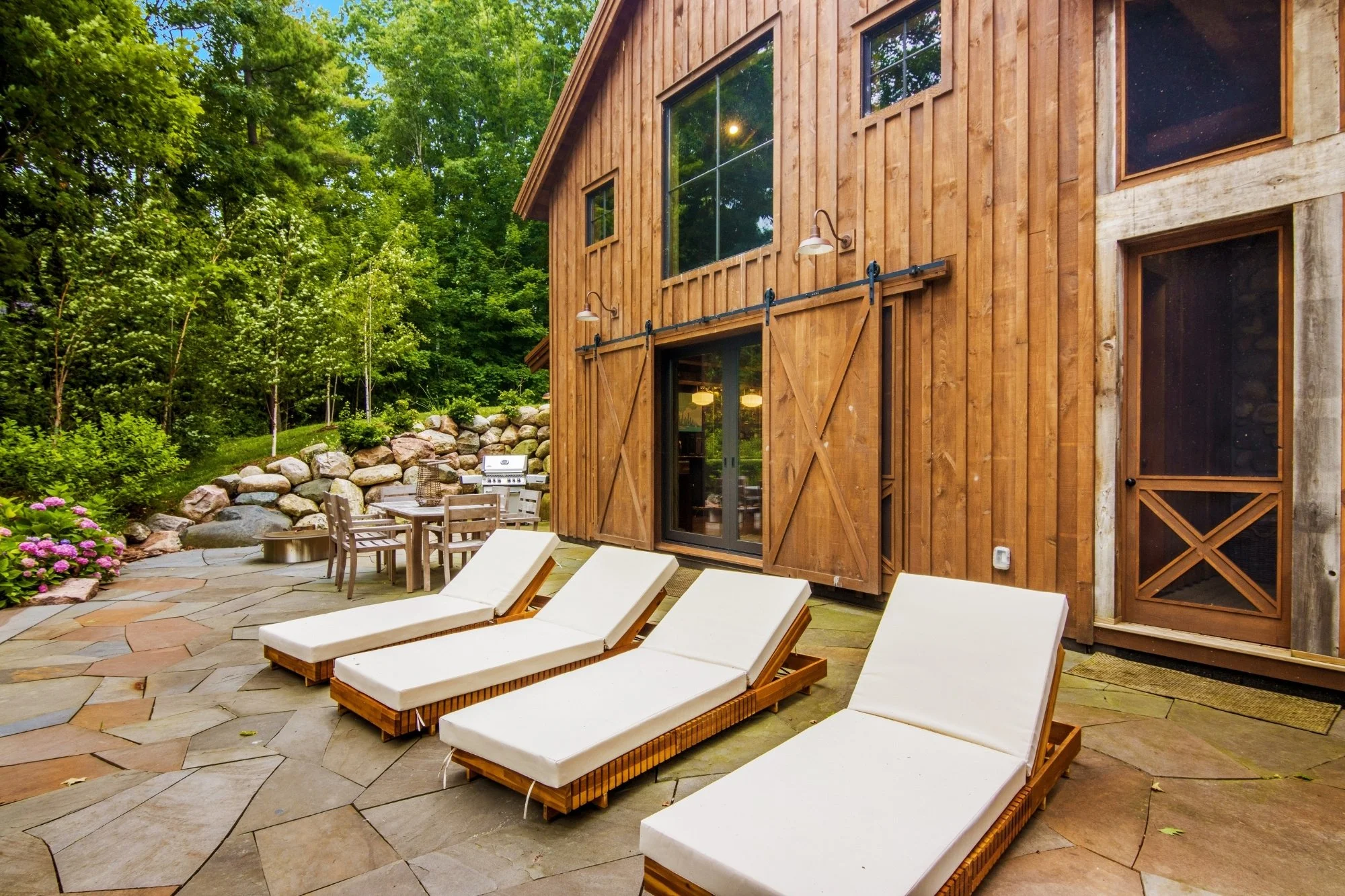A backyard patio with four white cushioned lounge chairs, a stone patio, and a wooden house with a sliding glass door, wood siding, and barn-style doors. There is a table with chairs and a grill, surrounded by greenery and trees.
