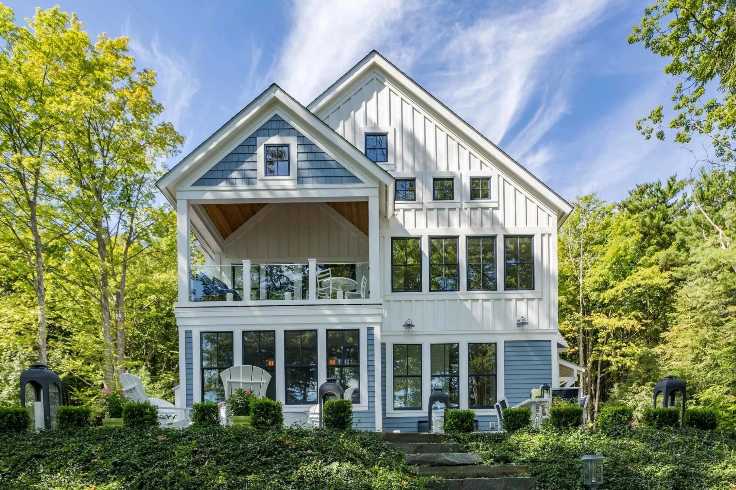A white modern house with blue accents features multiple windows and a balcony, surrounded by lush green trees on a clear sunny day with blue sky and wispy clouds.