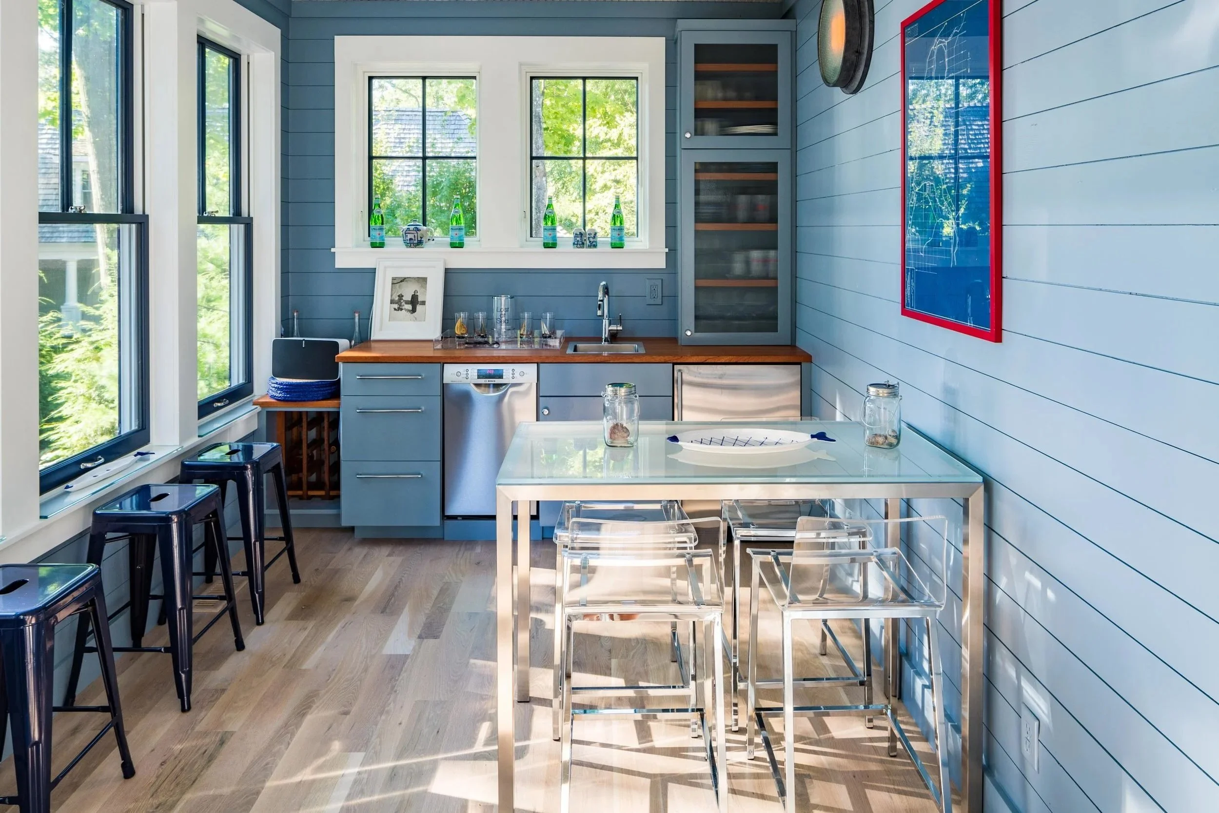 A bright kitchen with large windows, light blue shiplap walls, and wooden floors. There are black bar stools on the left, a white dining table with clear chairs in the center, and blue cabinets with glass doors. Decorative bottles and framed photos are on the windowsill, and a red-framed blue map hangs on the right wall.