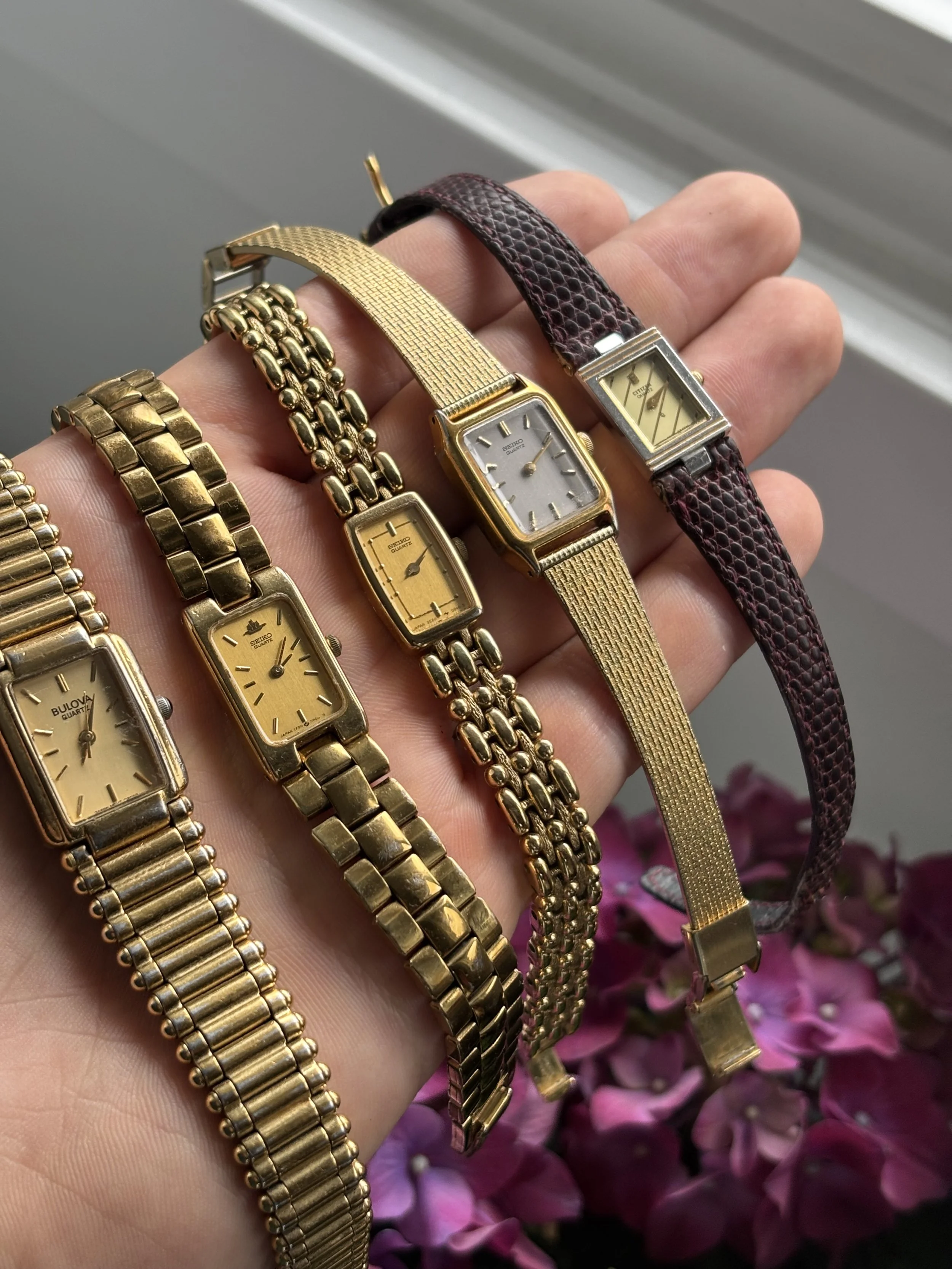 A person's hand holding several vintage gold-toned wristwatches and bracelets in front of pink flowers and a window.