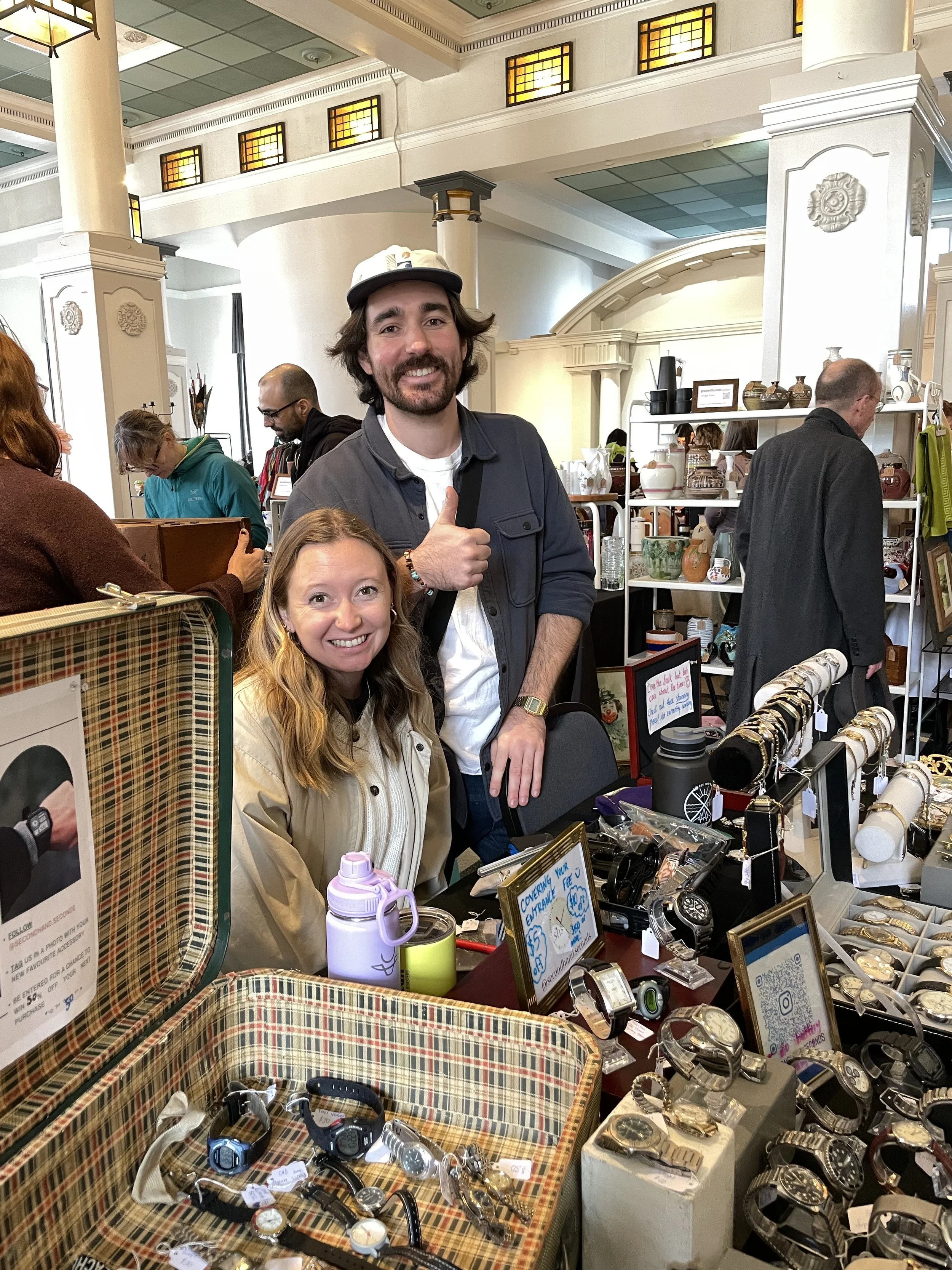 A man and a woman at a jewelry table with watches and accessories, in a store with shelves of pottery and other items in the background.