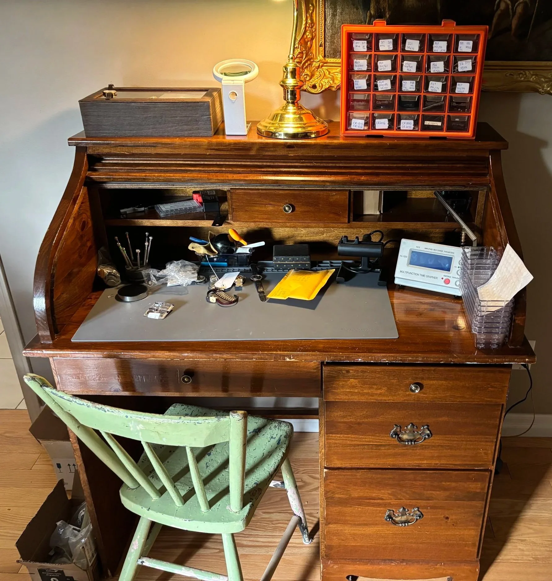 A cluttered wooden desk with various small objects, a green chair, and a white chair in front of it. The desk holds a multicolor multimeter, a box, loose small items, and a plastic container. The background features a wall with a gold-framed mirror, and items like storage boxes, a gold-colored lamp, and a red organizer with small labeled compartments.