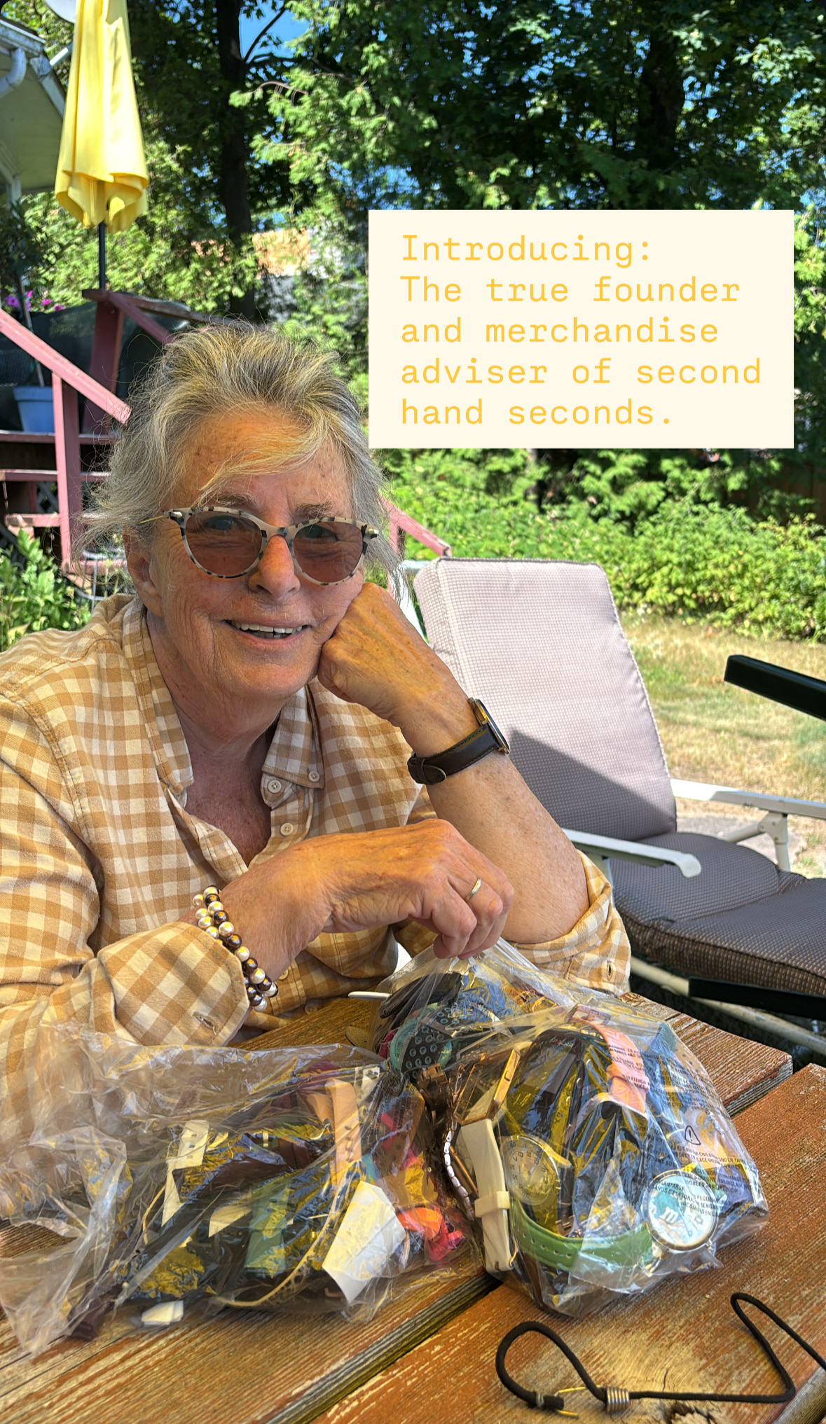 An elderly woman wearing glasses and a checkered shirt, sitting outdoors at a table with a bag of assorted secondhand sunglasses. There are trees in the background and a vacant chair nearby. A text overlay introduces her as the founder and merchandise adviser of second-hand seconds.