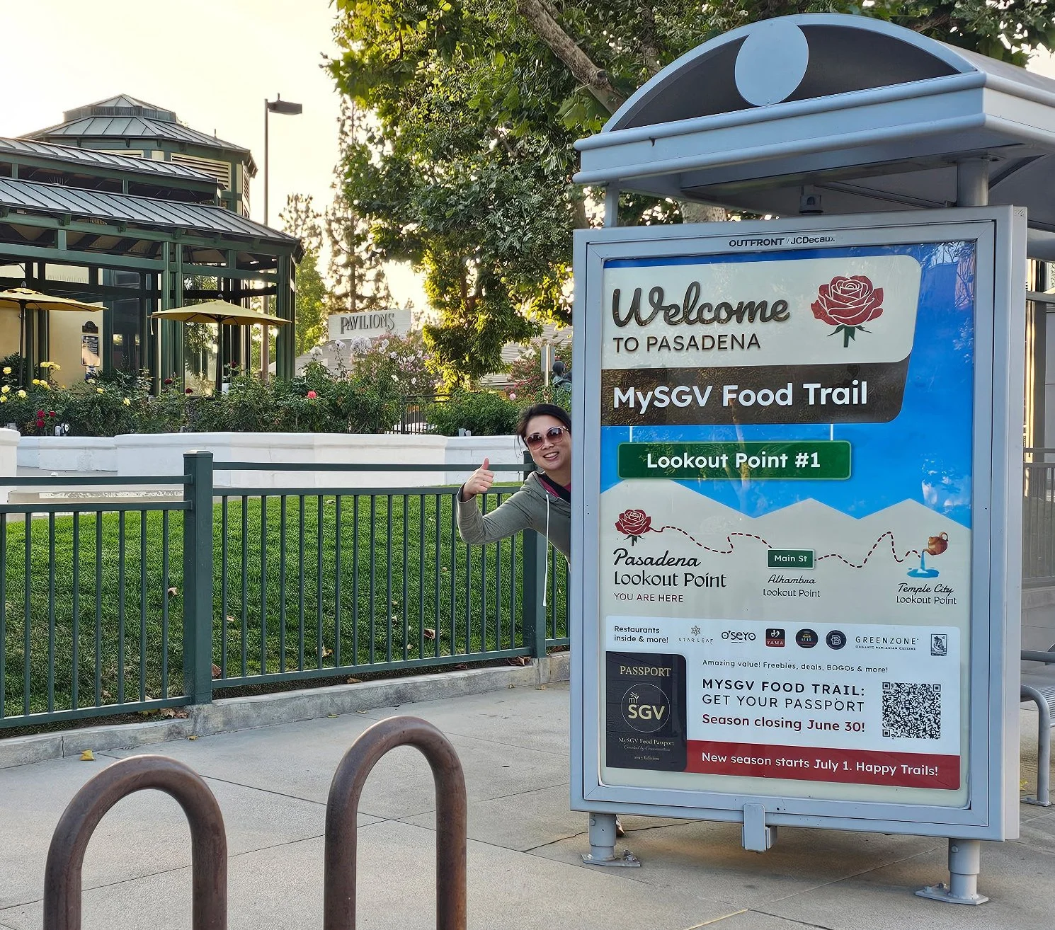 A person giving a thumbs up peeks from behind a large sign at a public park or event entryway. The sign reads 'Welcome to Pasadena MySGV Food Trail,' and has a map showing two lookout points, Pasadena and Temple City. The background features trees, a building with a green roof, and outdoor umbrellas.