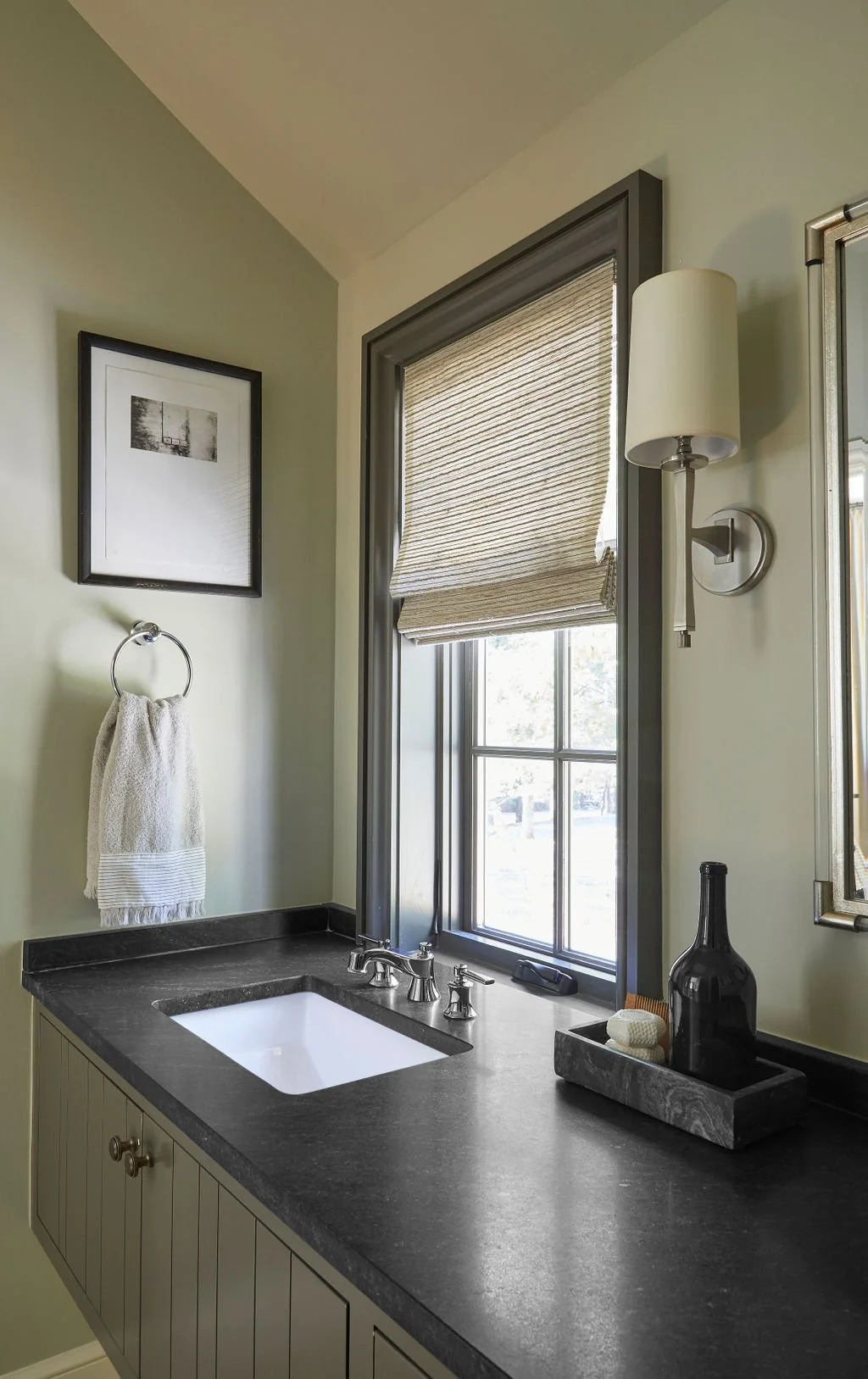 Bathroom with a dark countertop, a white undermount sink, a window with a woven shade, a wall-mounted light fixture, a towel ring with a towel, a framed black and white picture, and decorative items on a tray.
