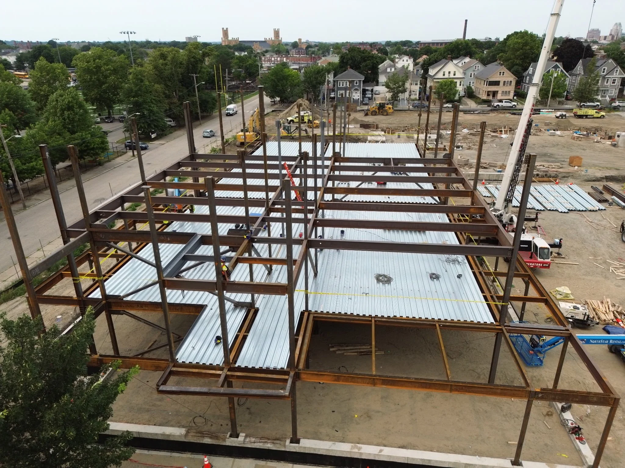 Construction site with steel framework, workers, construction equipment, and a crane in an urban area with residential houses and buildings in the background.