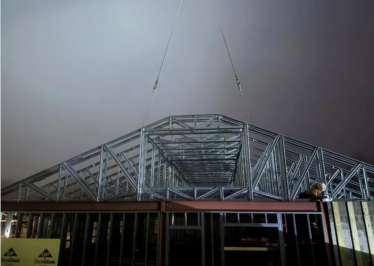 Construction worker in hard hat working on a metal framework of a building under construction, with a crane lifting part of the structure in an indoor setting.