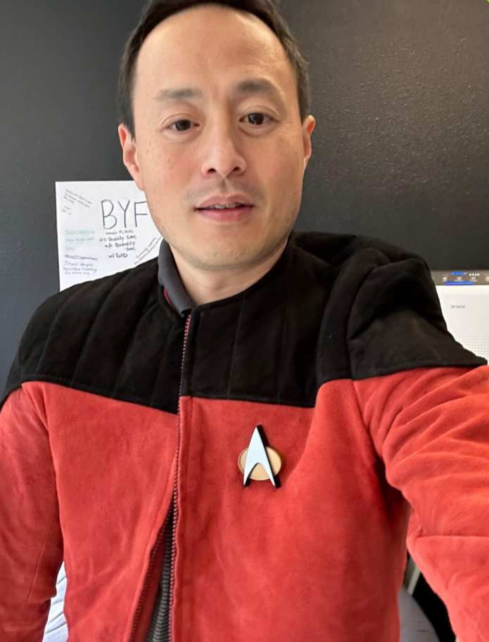 A man wearing a Star Trek uniform with a red and black jacket and the Starfleet emblem, taking a selfie in front of a dark wall with a paper that has 'BYE' written on it.