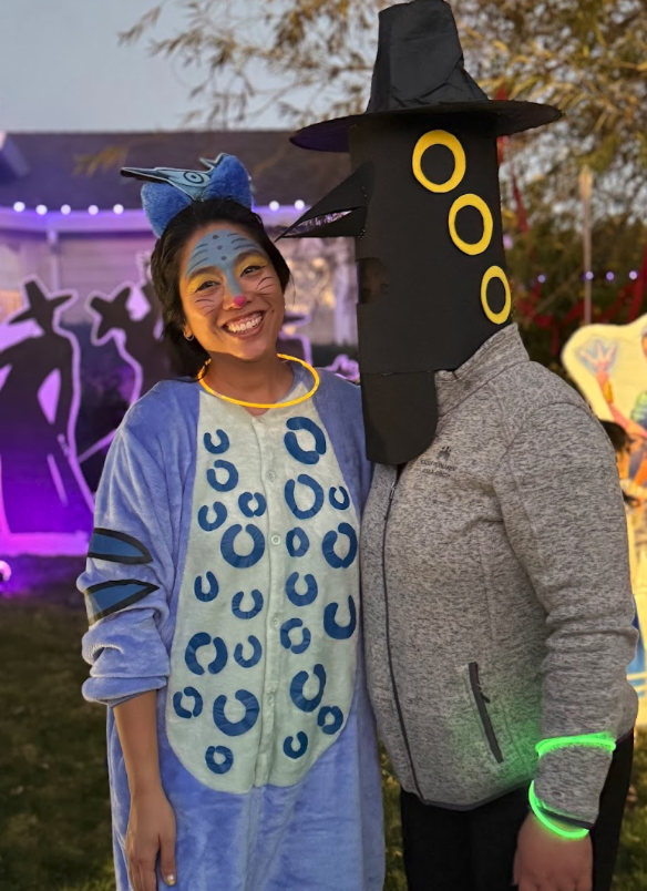 Two people dressed in costumes posing at an outdoor Halloween event. The person on the left is dressed as a colorful character with blue and yellow face paint, a blue costume with darker blue spots, and a headband with large blue and purple animal ears. The person on the right is wearing a black witch hat and a mask resembling a black cauldron with three yellow circles and purple stars.