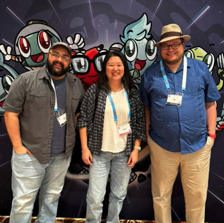 Three people standing in front of a colorful cartoon-themed backdrop at a convention.